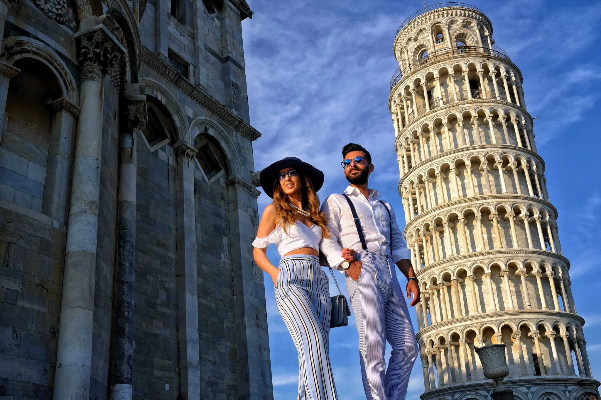 A young woman and man standing in front of the Leaning Tower of Pisa, with historic stone buildings nearby, during daylight.