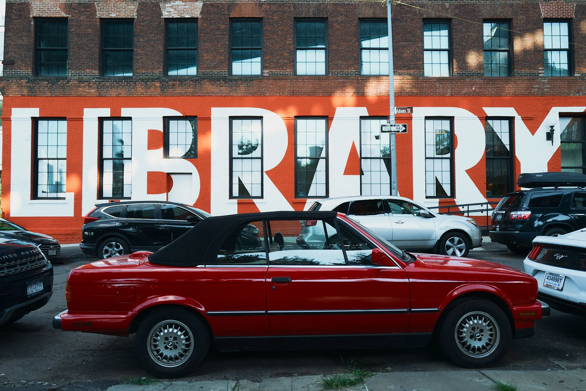A red convertible car parked on the street in front of a large building with a mural spelling out 'LIBRARY' on the side.