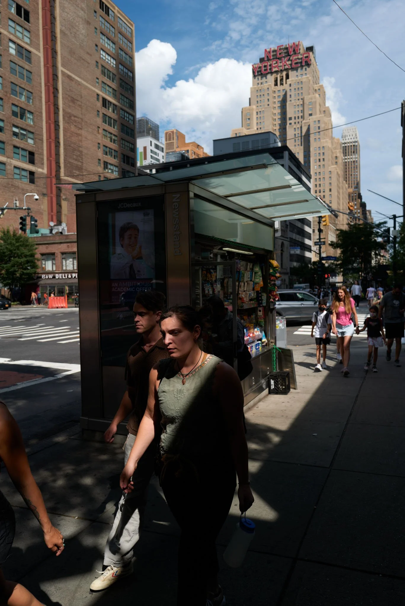 City street scene with pedestrians walking past a newsstand on a sunny day, with a tall building in the background displaying a neon sign that reads 'NEW YORKER'.