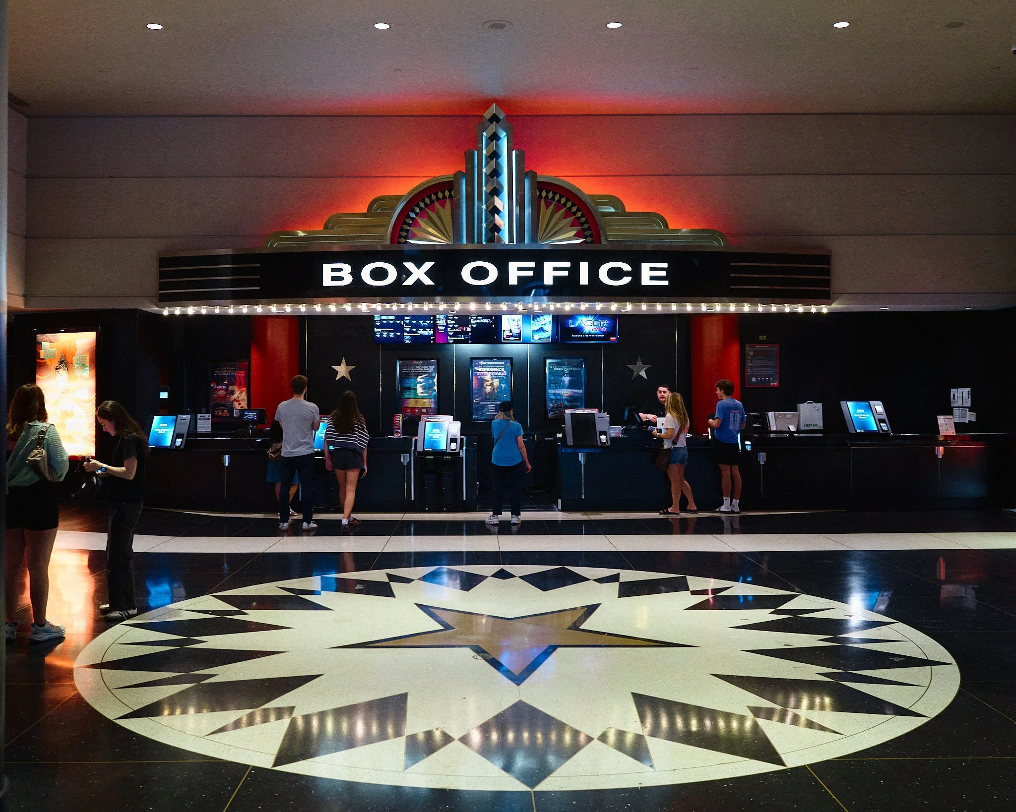 People buying tickets at a movie theater box office with an Art Deco design, illuminated sign, and movie posters visible.