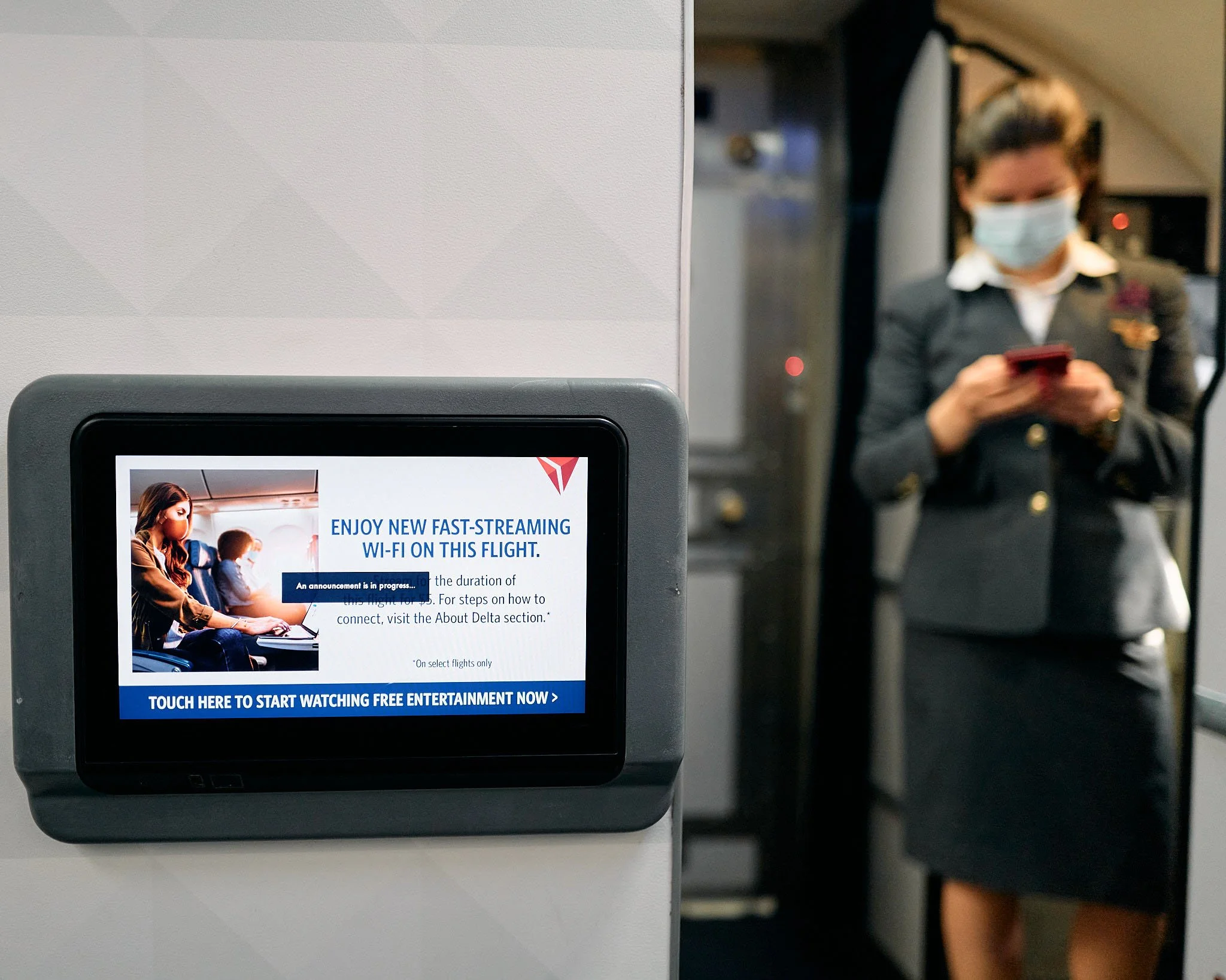 A person in a flight uniform wearing a face mask stands in front of a boarding gate, looking at their phone. A screen displays an advertisement for free Wi-Fi on the flight, with the Delta logo and a picture of passengers on an airplane.