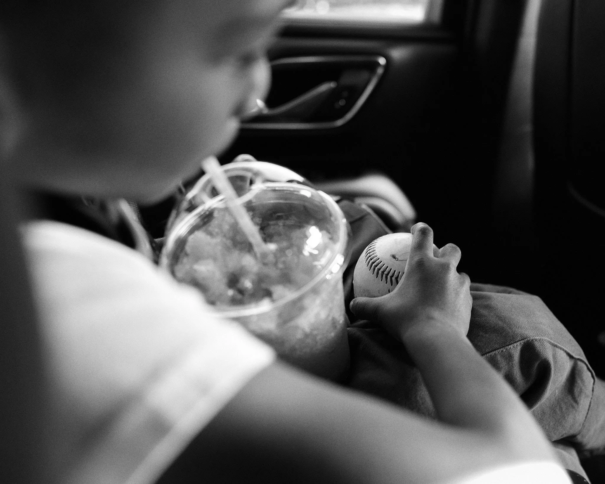 A child sitting in a car holding a baseball with one hand and drinking from a cup with a straw with the other hand. The photo is in black and white.