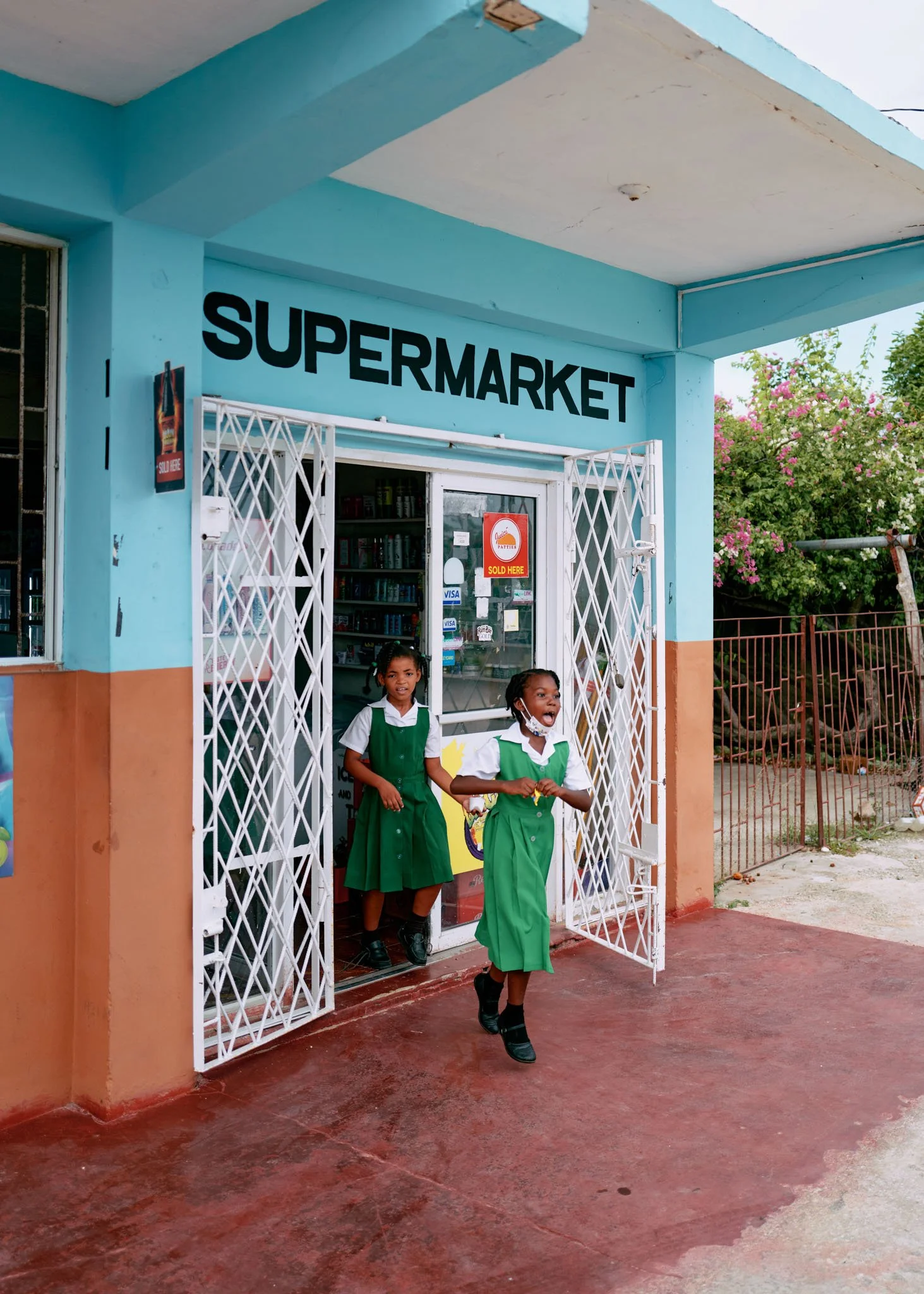 Two girls in green and white school uniforms leaving a small supermarket, with one girl stepping out and the other standing inside. The store has a blue exterior with the sign 'SUPERMARKET' above the entrance, and a partly open door with a security g
