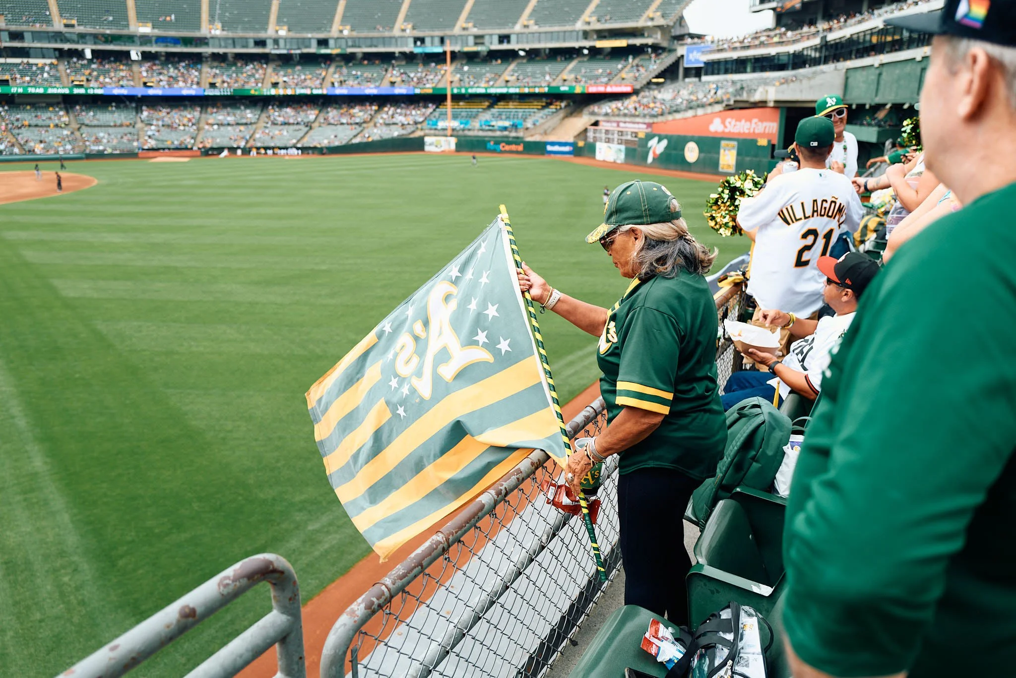 A woman in a green and yellow baseball jersey and cap is standing near the outfield fence, holding a scarf with the Oakland Athletics logo and flags, during a baseball game at a stadium.