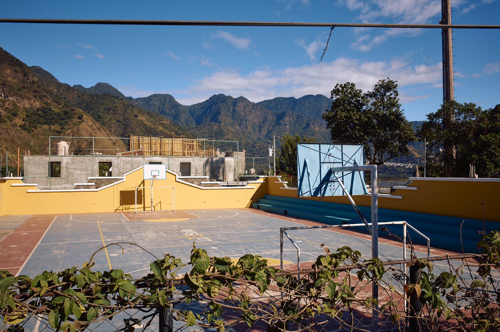 Empty outdoor basketball court with yellow walls, two hoops, and a blue scoreboard, surrounded by a fence and set against mountainous terrain and a clear blue sky.