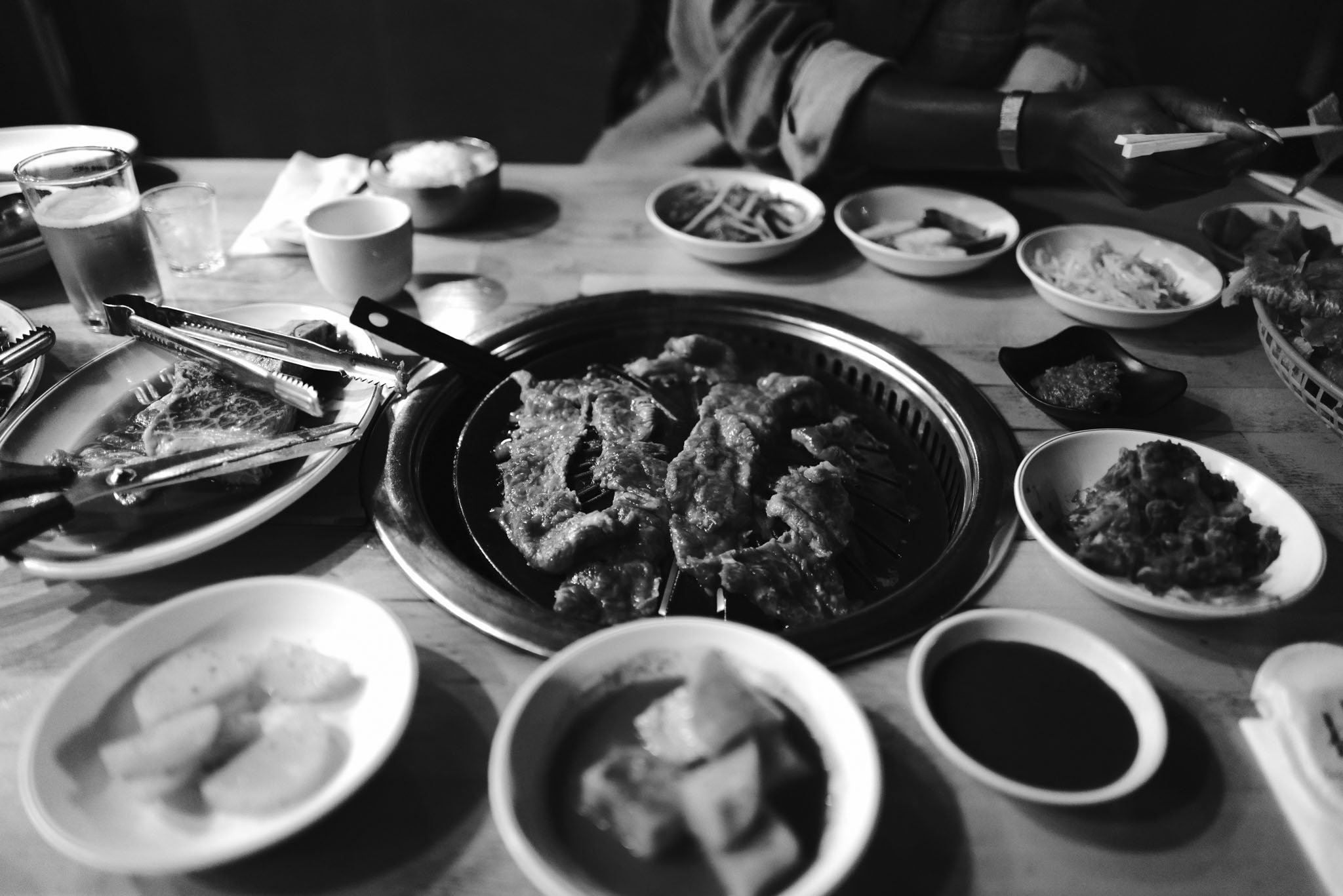 Black and white photo of a Korean barbecue table with grilled meat, various side dishes, sauces, and drinks, with a person using tongs to cook meat.