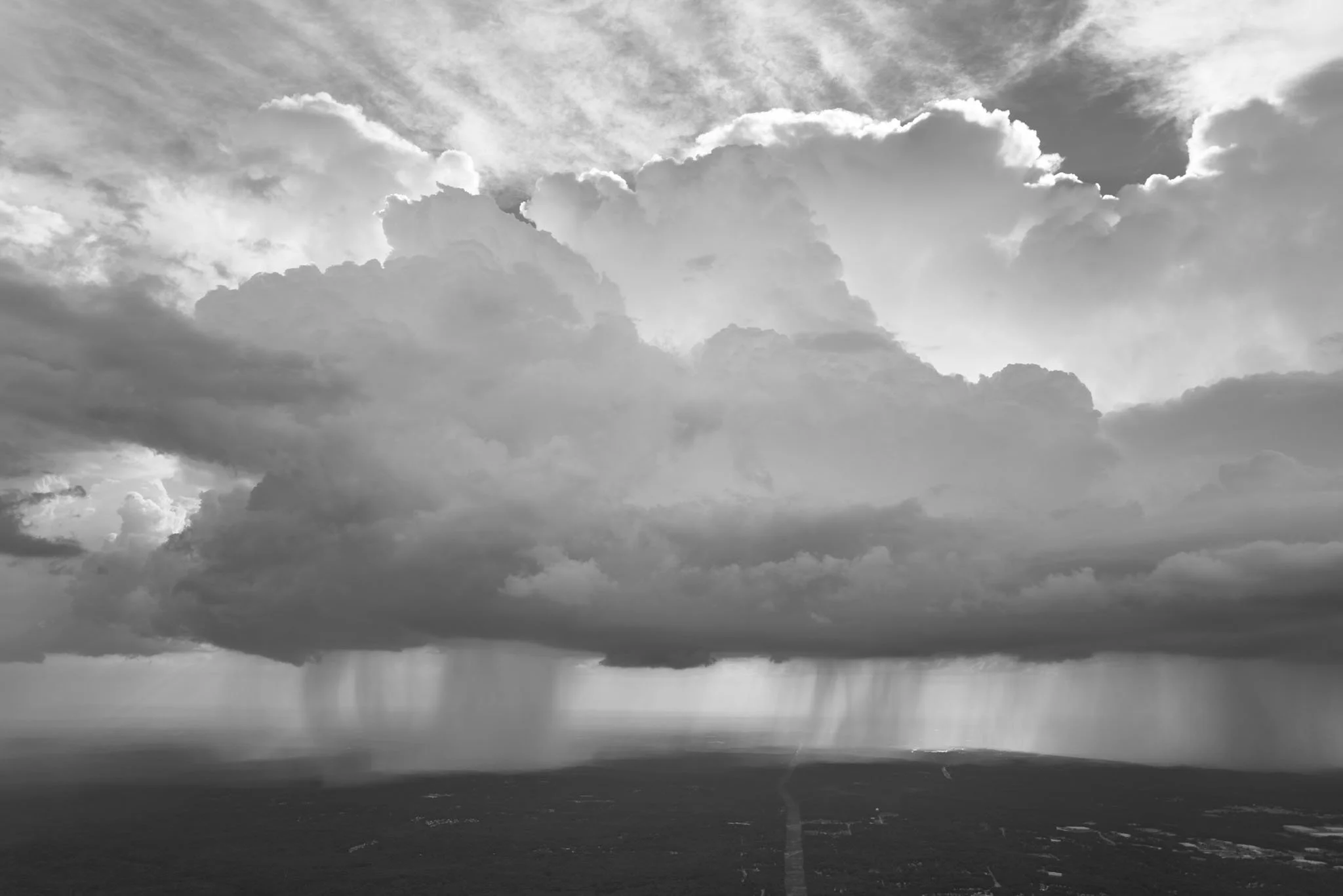 Black and white photo of a storm cloud with rain falling from it over a landscape with a distant horizon.