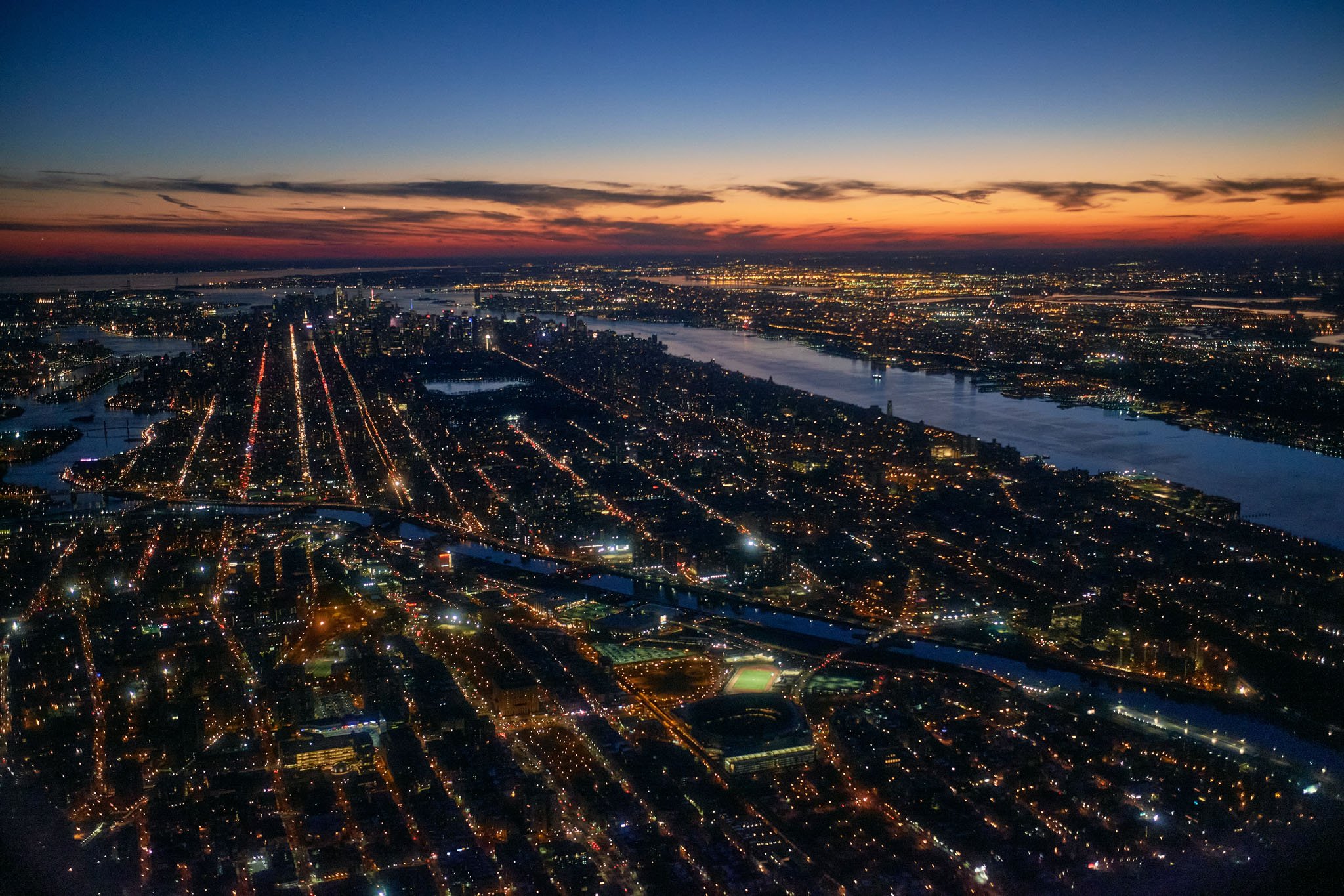 Aerial view of New York City at dusk with city lights, rivers, and a colorful sunset