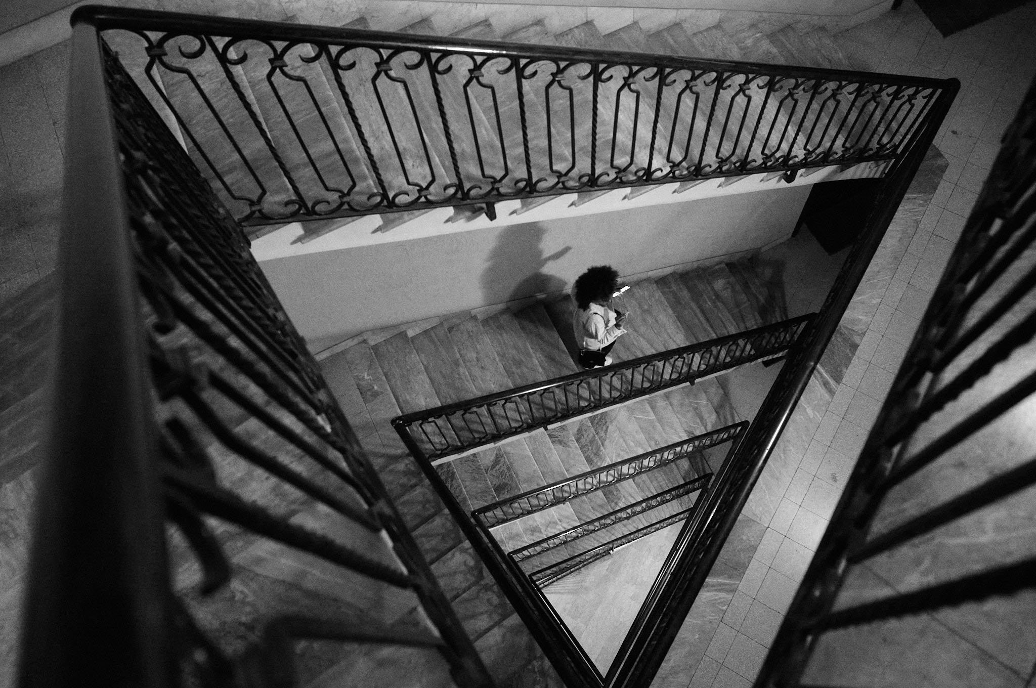 A black and white view looking down a spiral staircase with ornate railing, showing a woman with curly hair standing on a landing, looking at her phone.