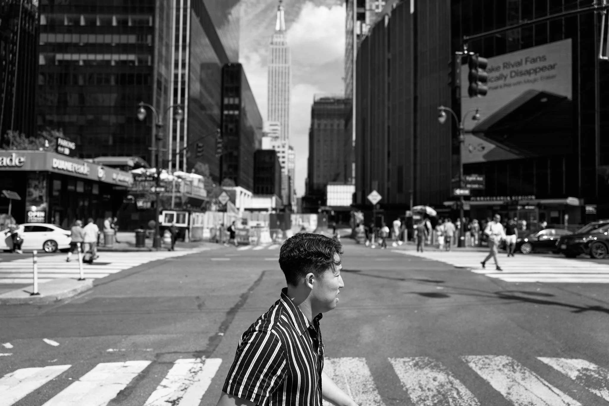 A young man wearing a striped shirt walking across a city street with skyscrapers in the background, black and white photo.