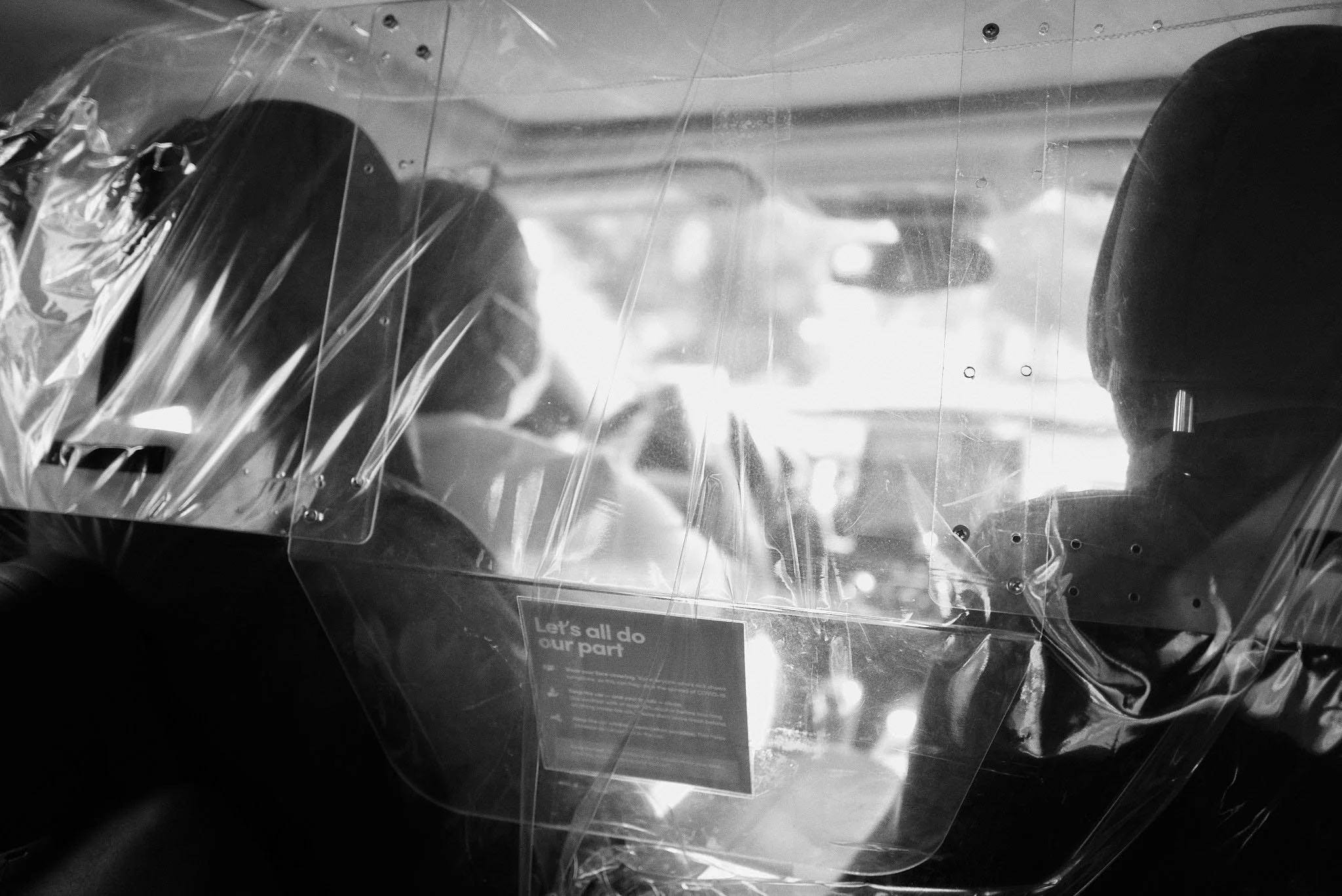 Two people sitting inside a vehicle with a clear partition between them, one of whom is wearing a mask, and a sign on the partition that reads "Let's all do our part."