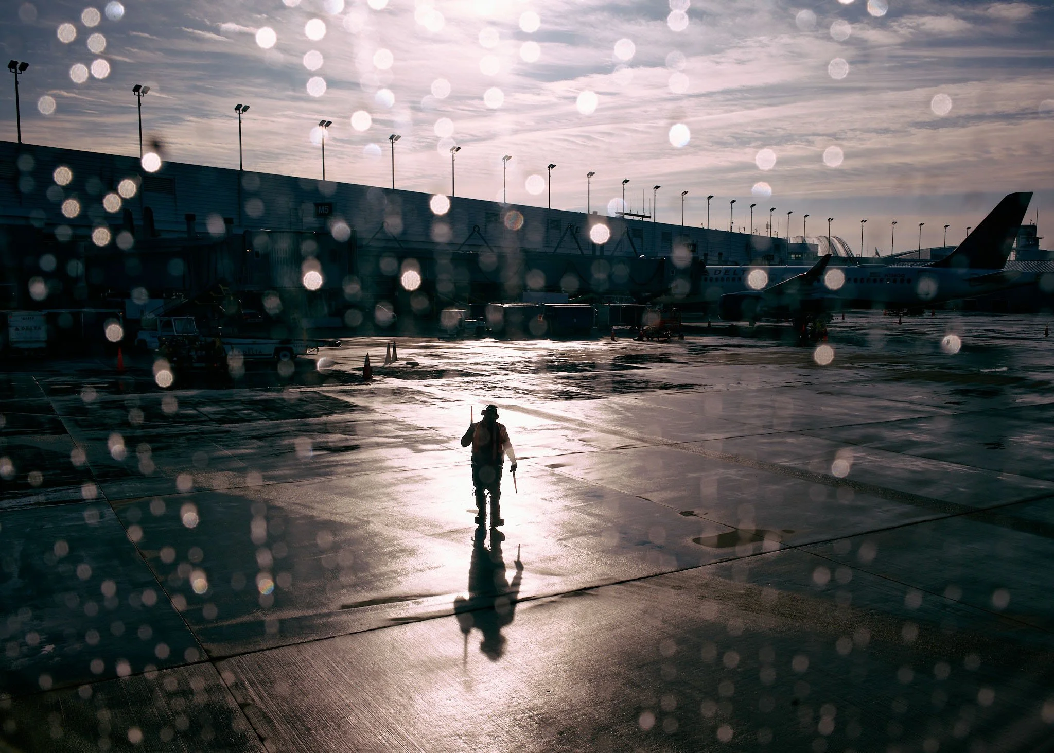 A person walking on a wet airport tarmac with airplanes parked at the gates in the background, viewed through a rain-spattered window during sunset or sunrise.
