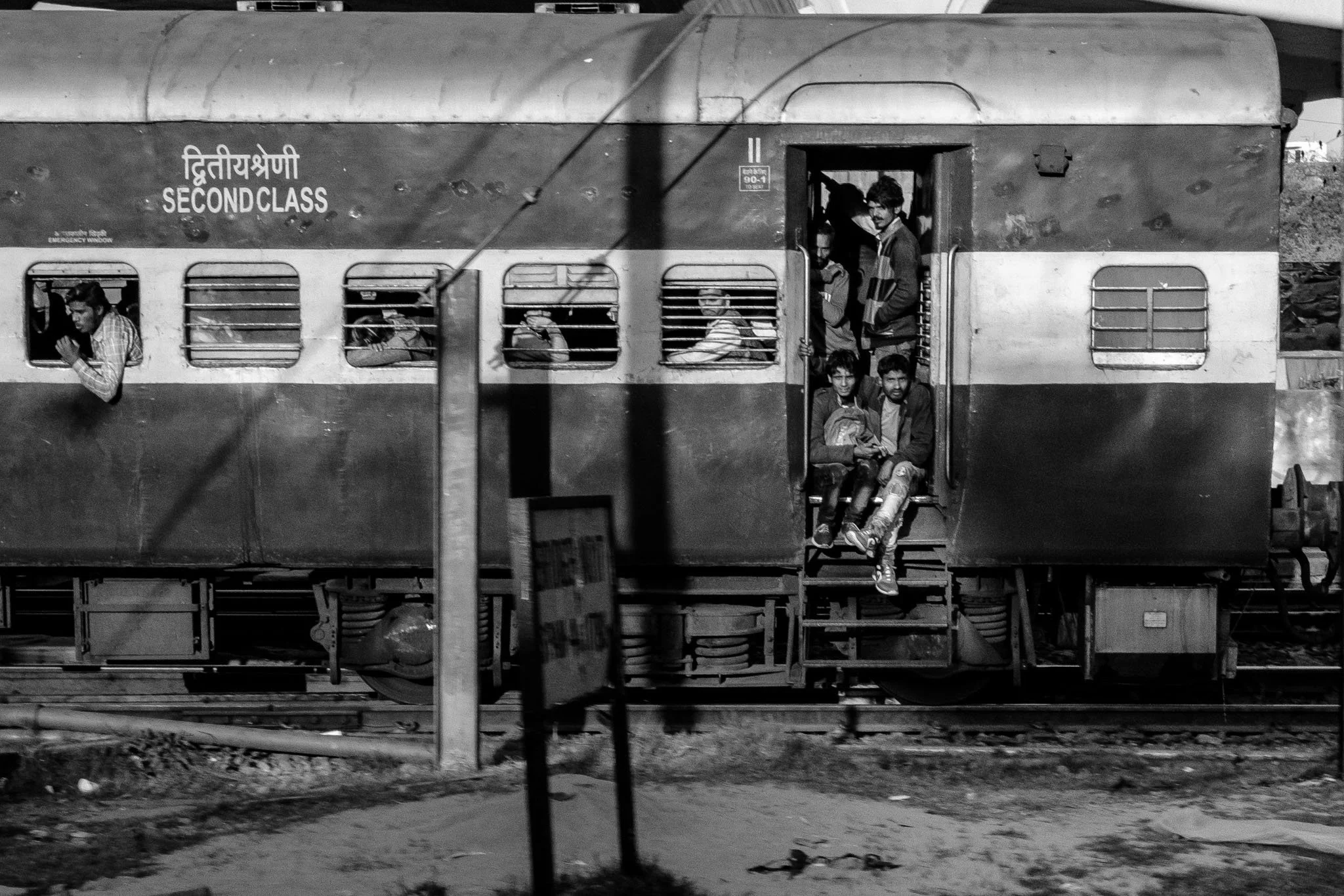 A black and white photo of a train with passengers, some looking out the windows and some sitting and standing in the doorway, on the station tracks.