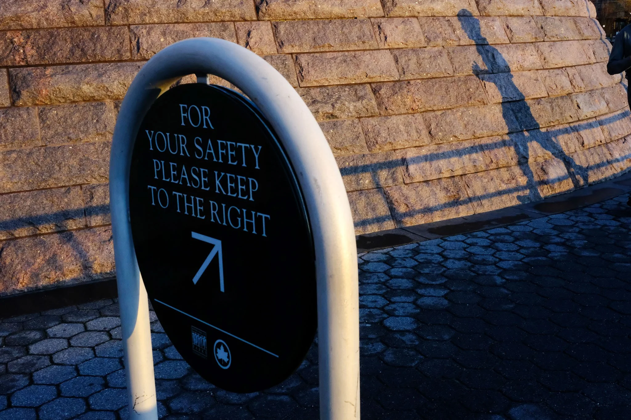 A black circular sign with white text that reads, 'For your safety please keep to the right,' attached to a white metal pole. The sign is situated on a cobblestone pavement with a brick wall in the background. The scene is illuminated by sunlight, ca