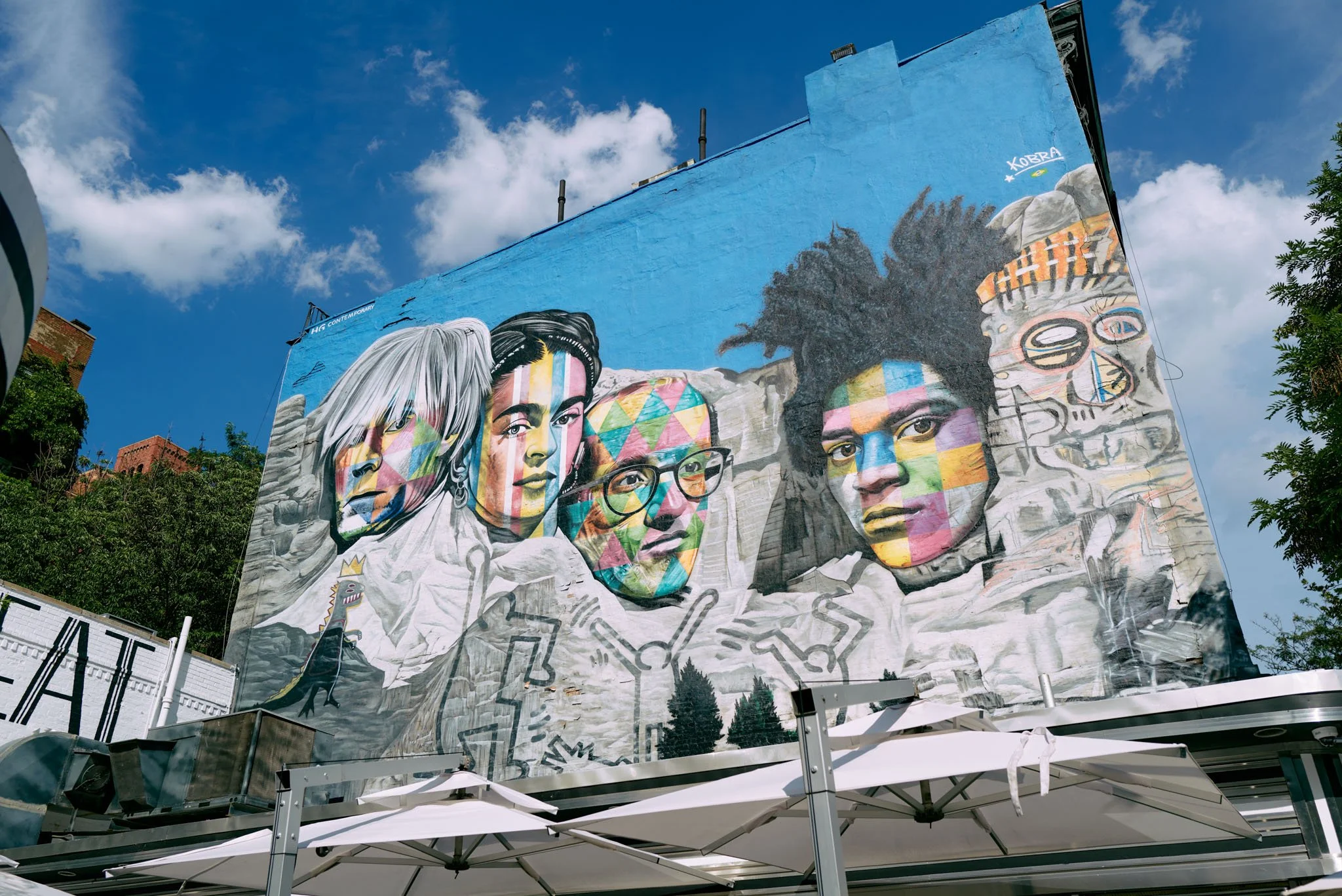 Colorful mural of four diverse women with face paint and glasses on a building wall, with a blue sky and trees in the background.