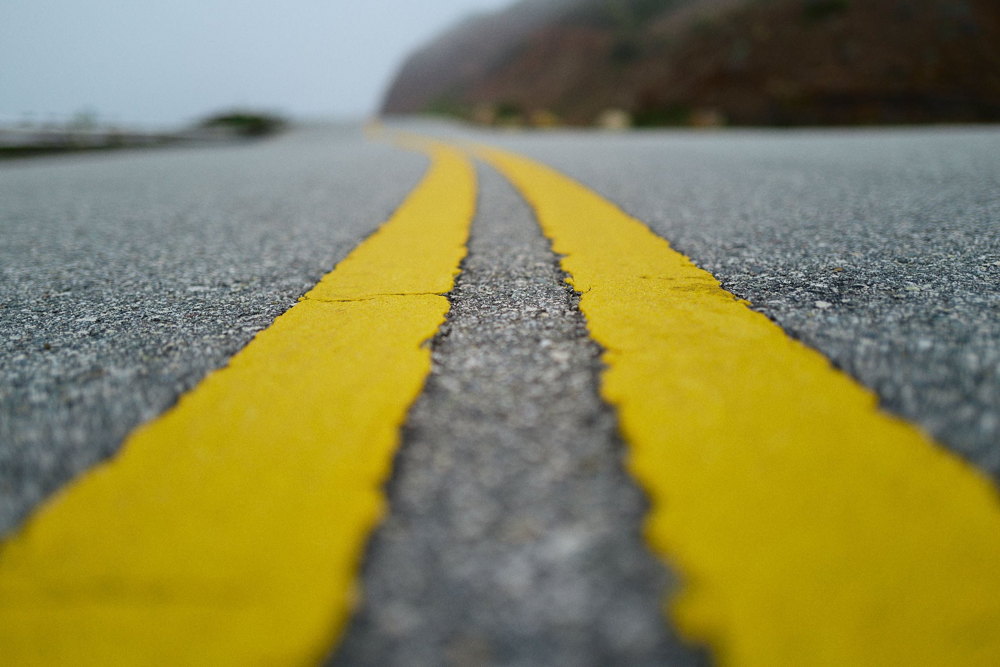 Close-up view of yellow double lines on a road surface, with a blurred background of a foggy landscape and distant hills.