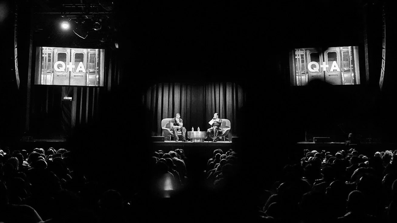 Black and white photo of a stage with two people seated in conversation, audience watching, with screens displaying 'Q+A' behind them. Chandeliers hang from the ceiling.