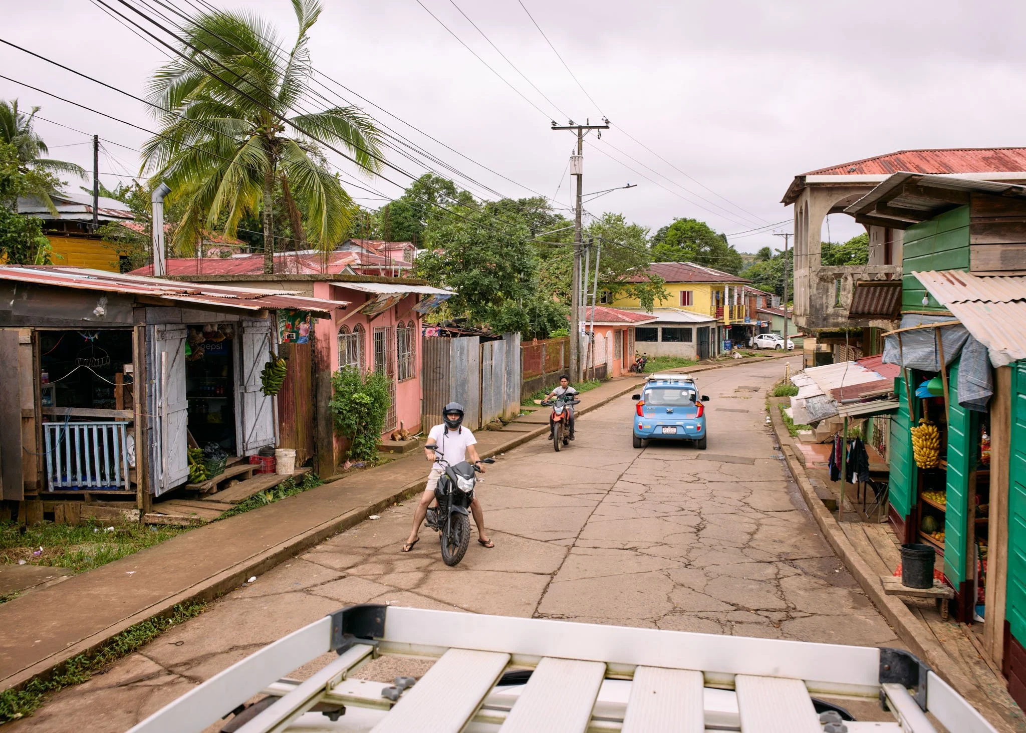 A residential street with small houses, some made of wood and others concrete, featuring colorful facades in pink, yellow, and green. The street is cracked and slightly curved, with utility poles and power lines overhead. There are two motorcyclists,