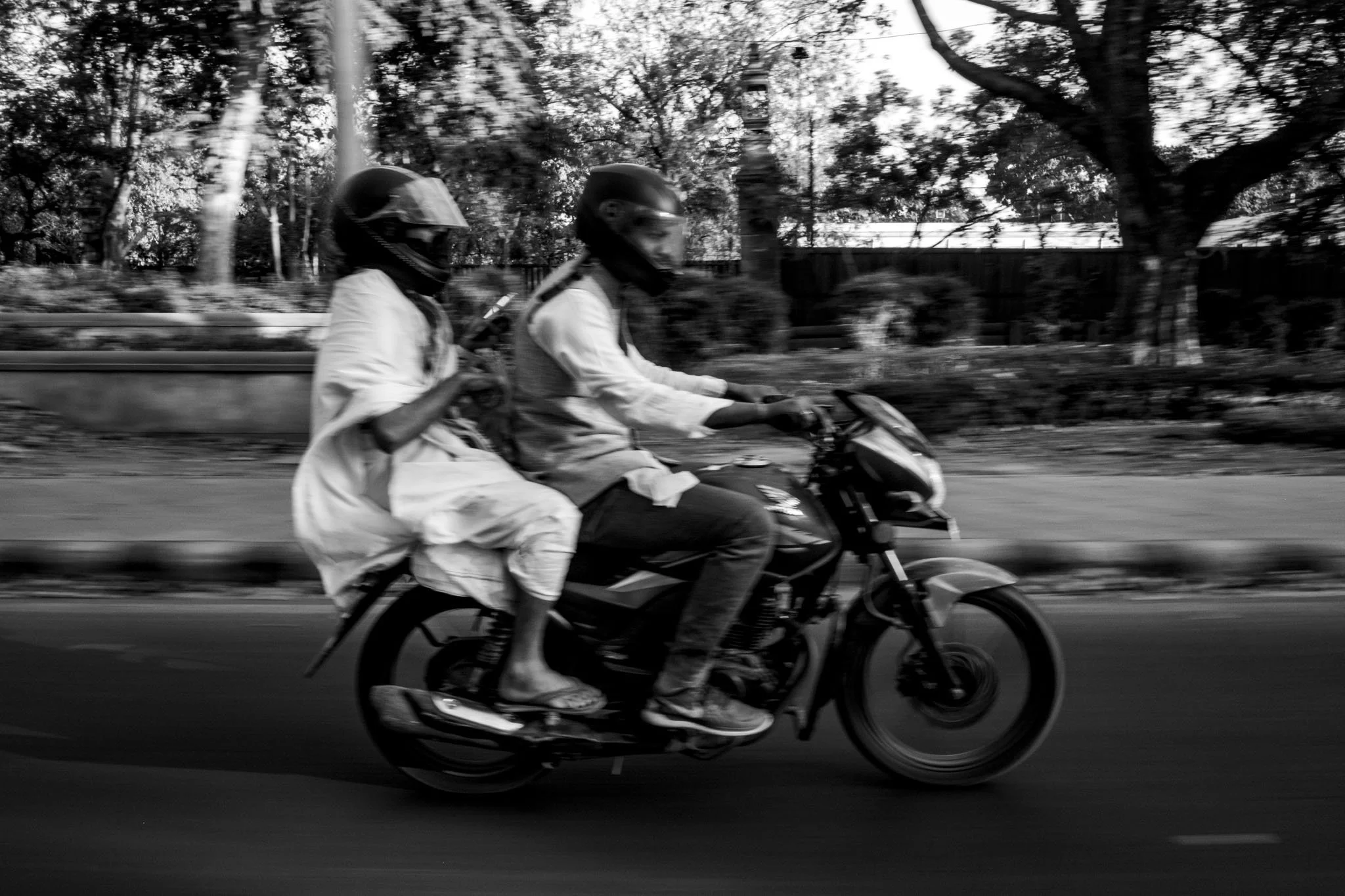 Two men wearing helmets riding a motorcycle on a street with trees and a fence in the background, black and white photo