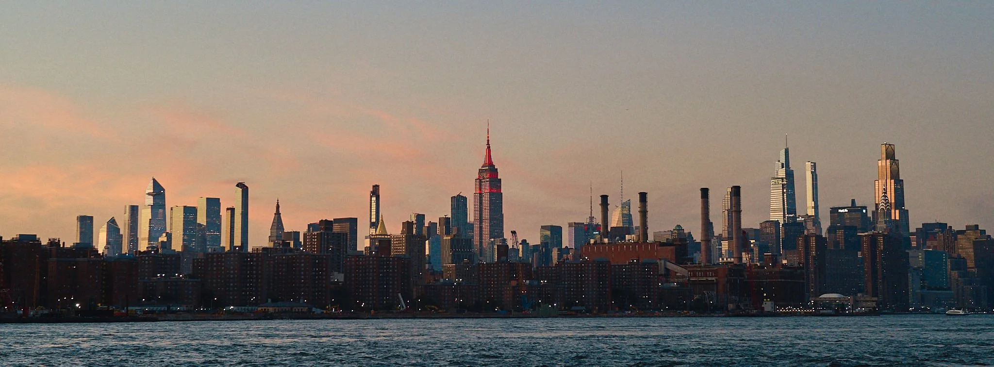 City skyline of Manhattan, New York City, with tall skyscrapers, including the Empire State Building, viewed from across the water at sunset.