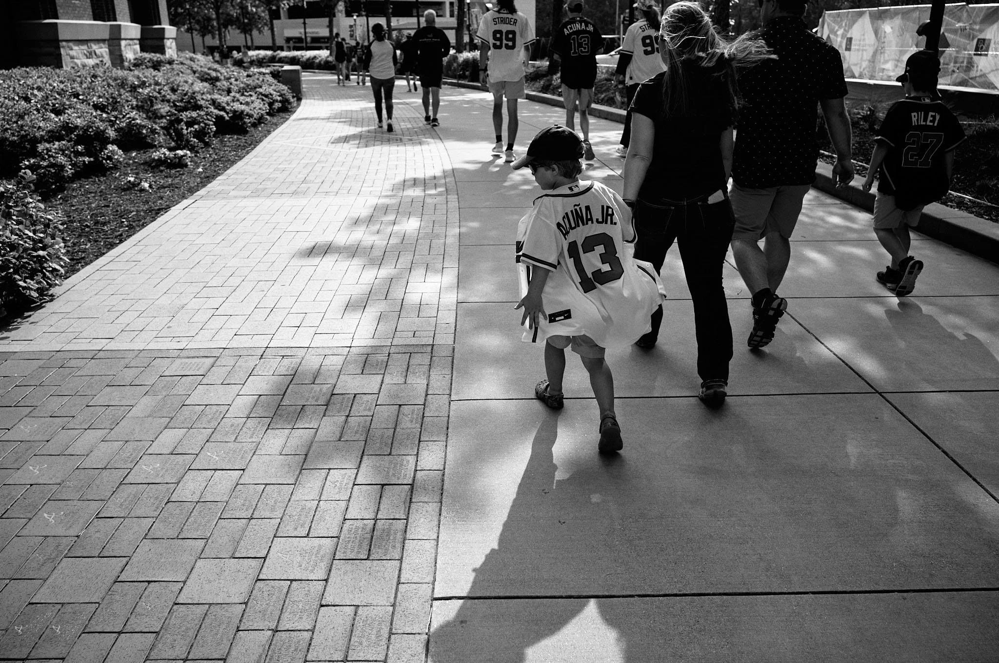 Children and adults walking on a sidewalk in a park-like setting, some wearing sports jerseys and casual clothing.