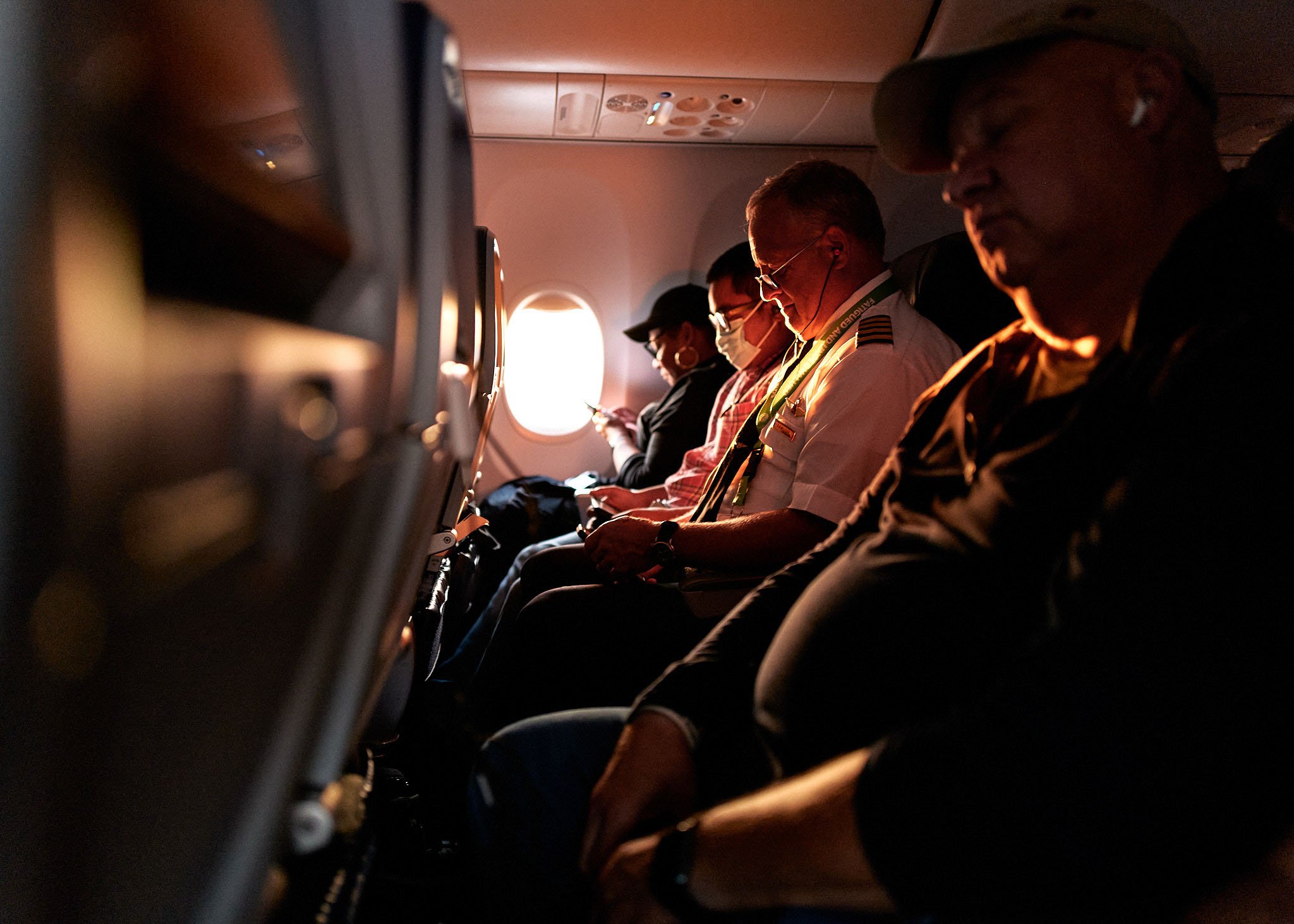 Passengers sitting in airplane seats with sunlight coming through the window, some wearing masks and others using electronic devices.