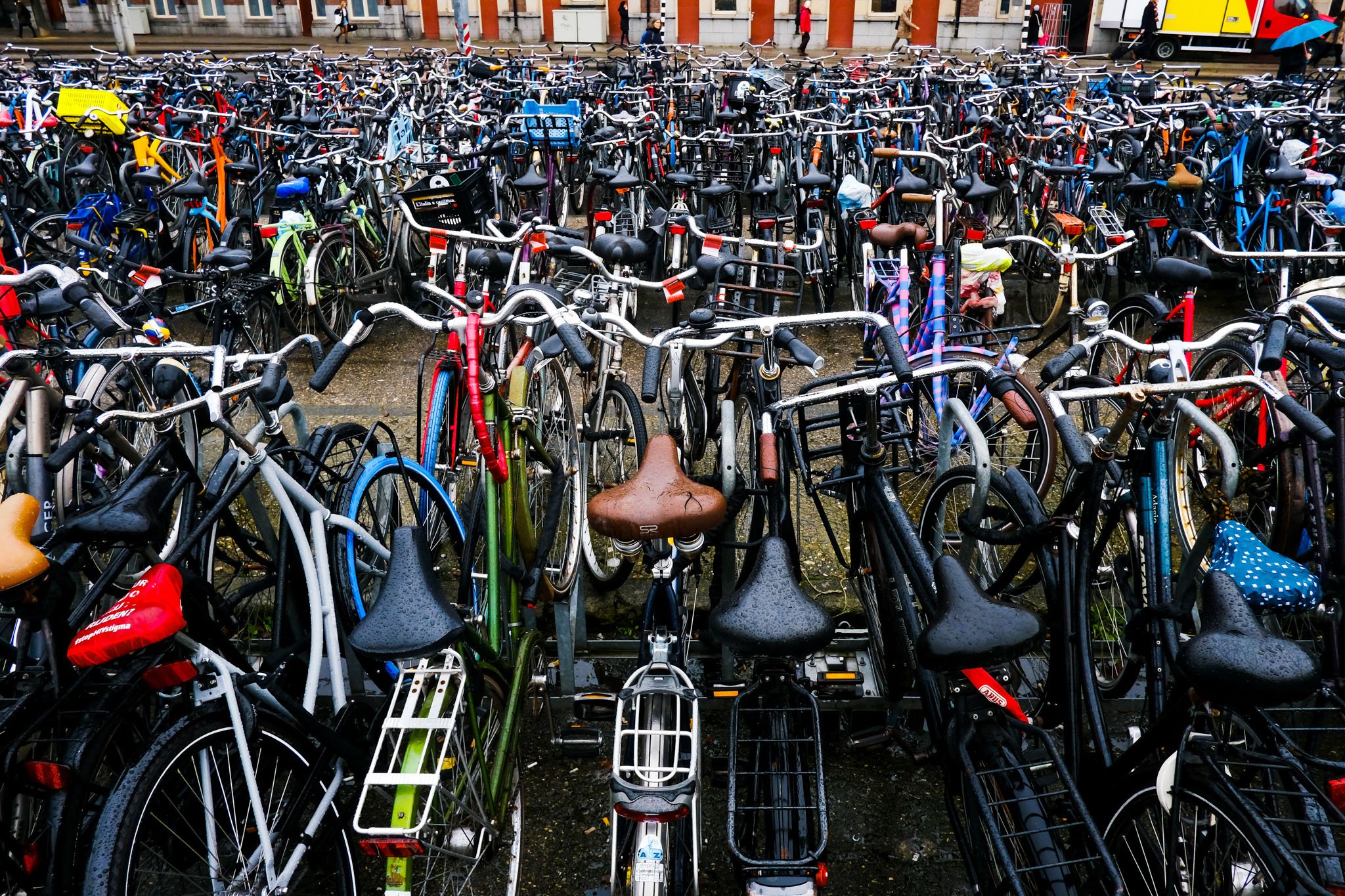 A densely packed bicycle parking lot with numerous bikes in various colors, styles, and sizes, parked tightly against each other on a wet surface, with buildings and a tram in the background.