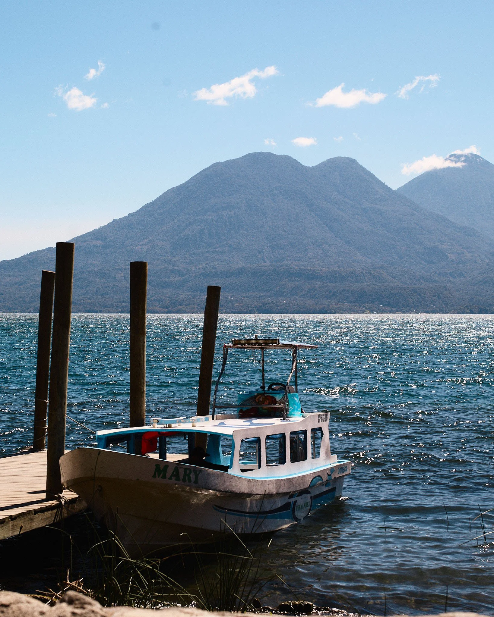Small white boat named 'Mary' docked at a wooden pier on a lake, with mountains in the background and a cloudy blue sky.