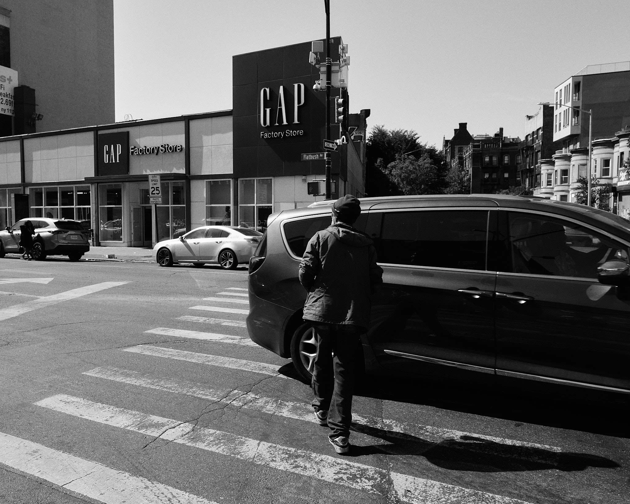 A person crossing the street at a crosswalk with a big black car in the foreground. In the background, there is a GAP factory store and other buildings.