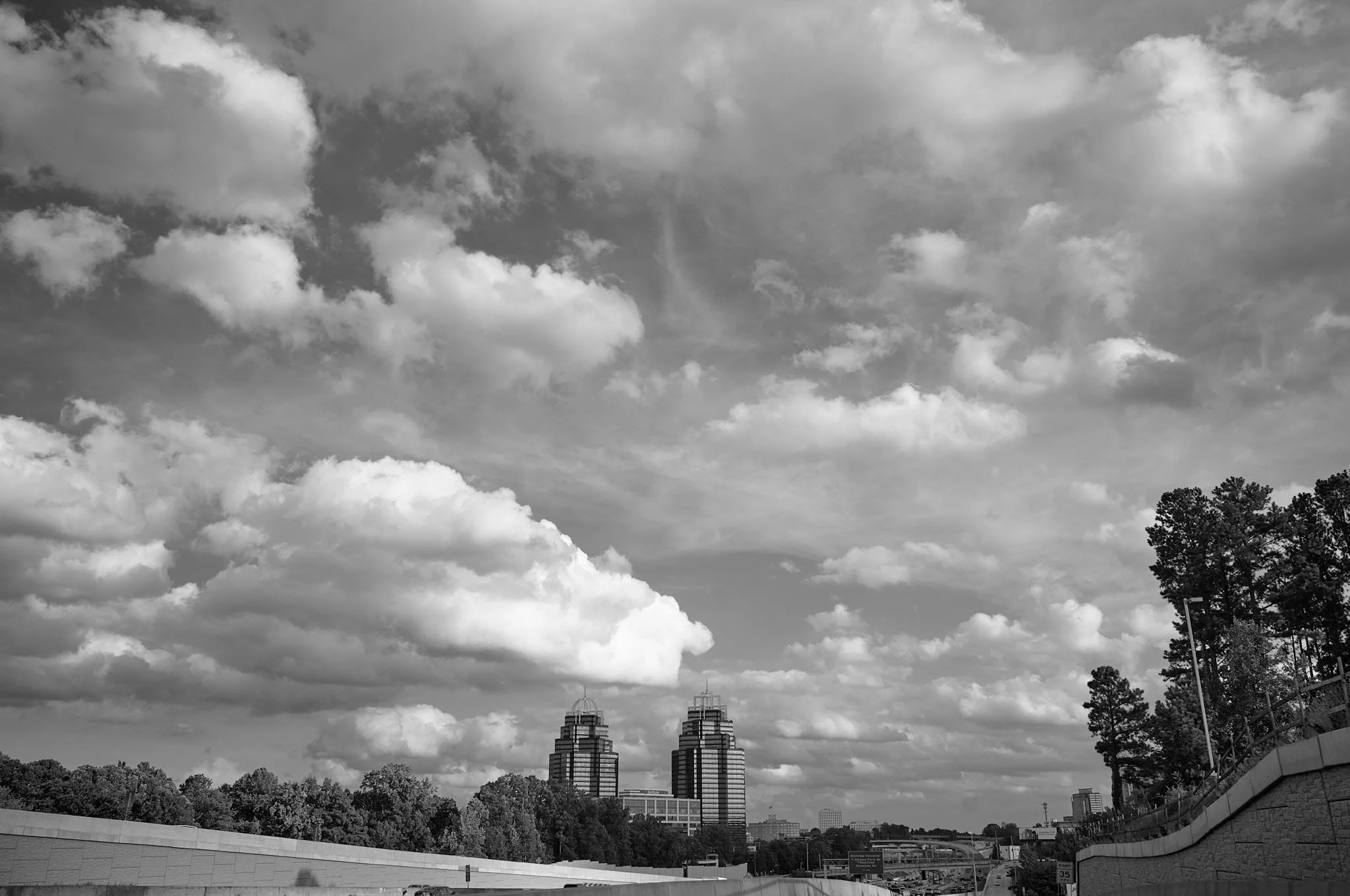 Black and white photo of a cityscape with two tall buildings, trees, and a partly cloudy sky.