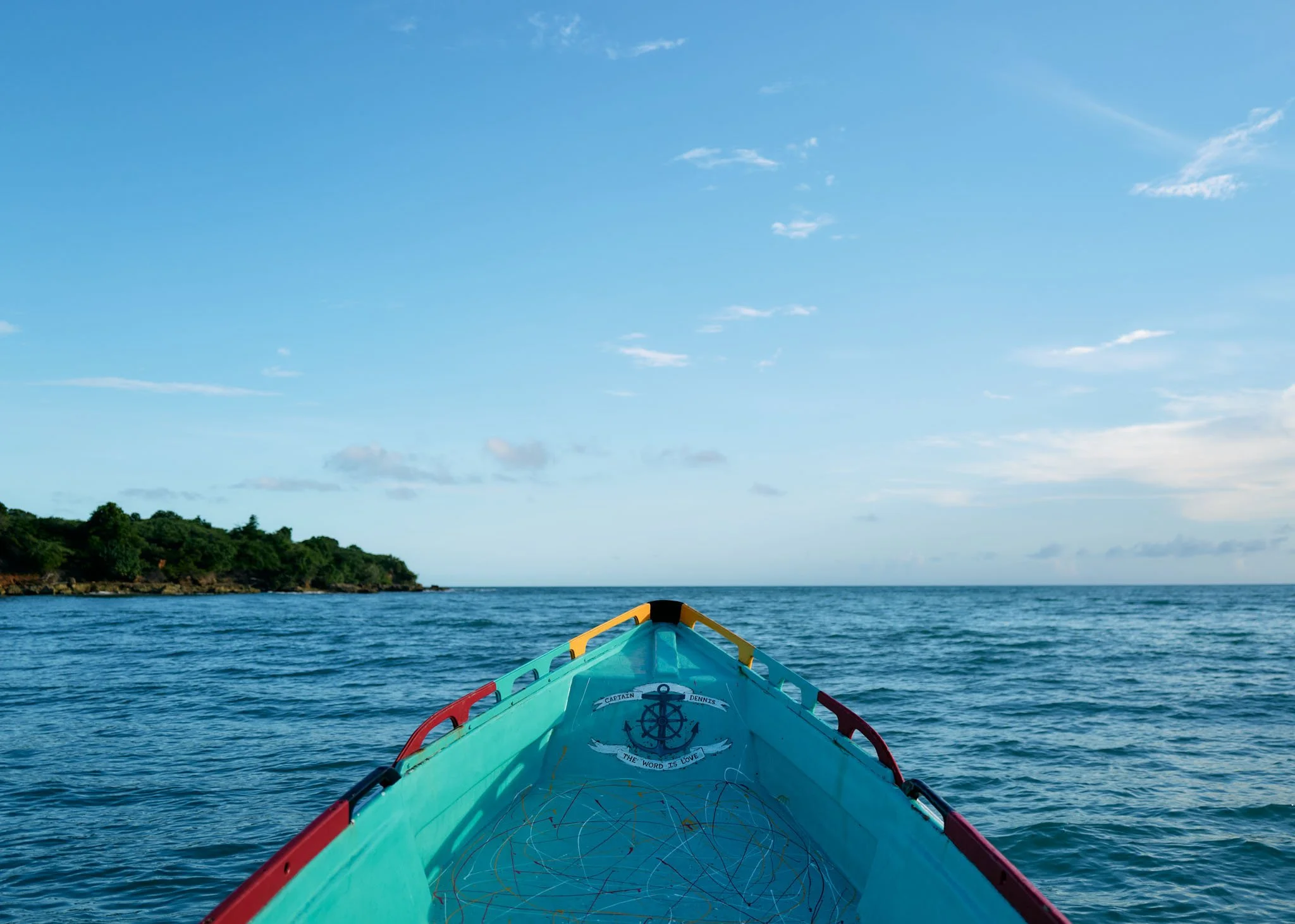 View from the front of a colorful boat with an anchor symbol, looking out on calm ocean waters with a small green island in the distance under a blue sky with scattered clouds.
