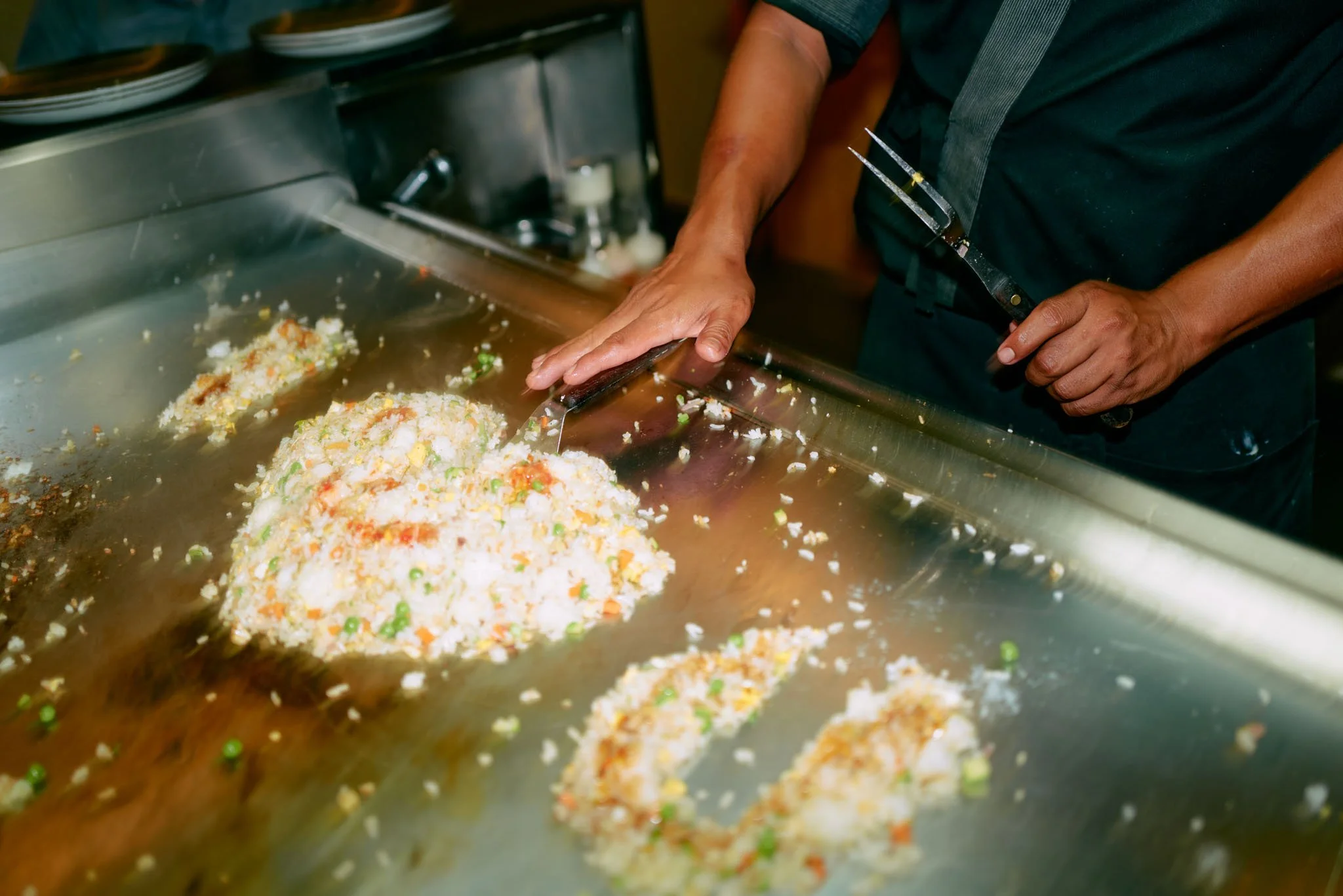 A person cooking fried rice on a large metal griddle, using a spatula to shape and stir the rice.