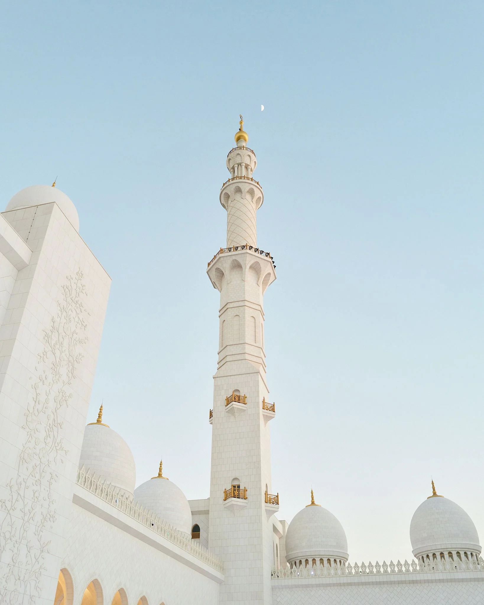 View of the Sheikh Zayed Grand Mosque's white minaret against a clear sky with a crescent moon.