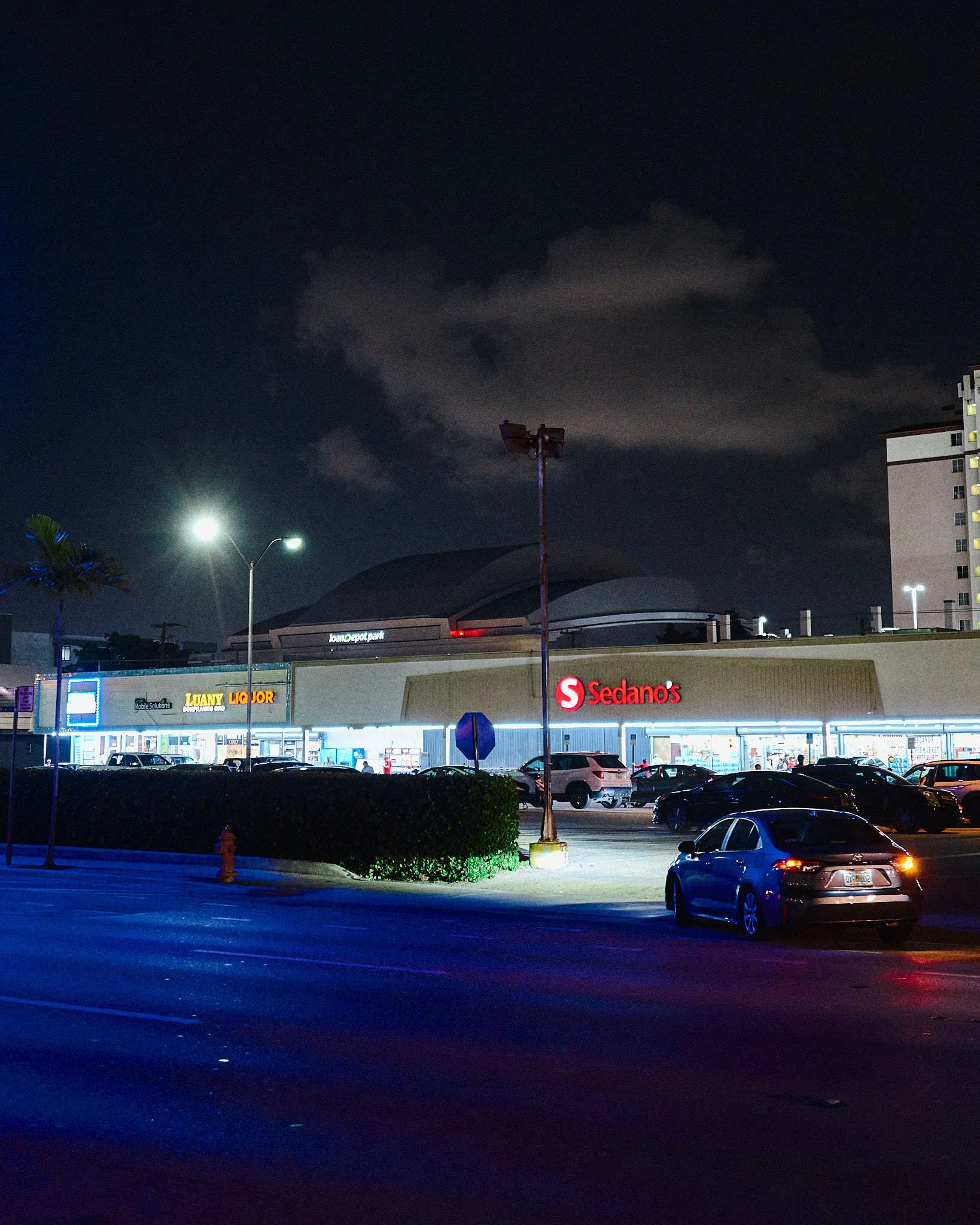 Night view of a shopping center parking lot with stores, parked cars, and streetlights, under a dark sky with clouds.