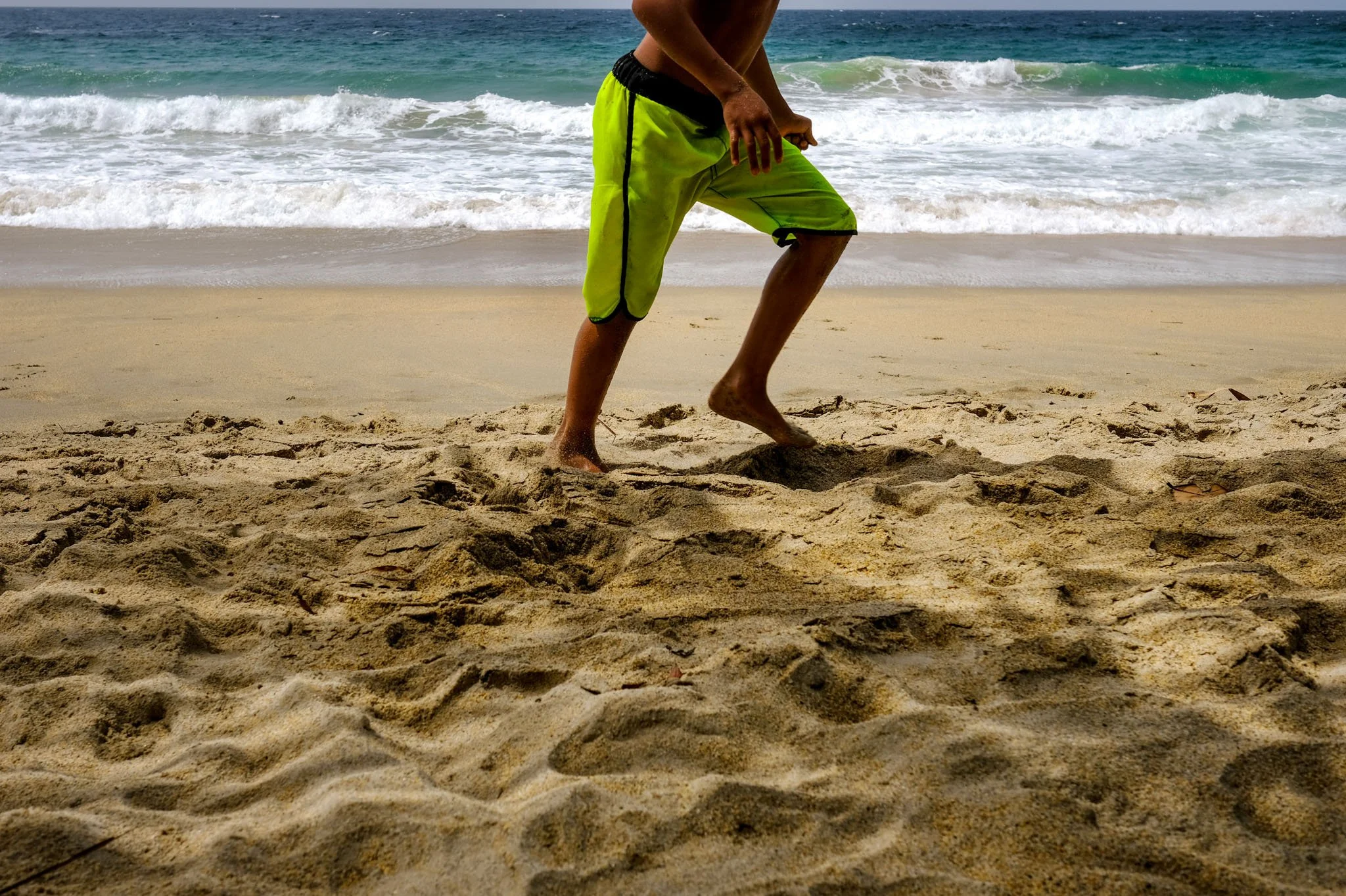 Person wearing neon green swim trunks walking along sandy beach with ocean waves in the background.