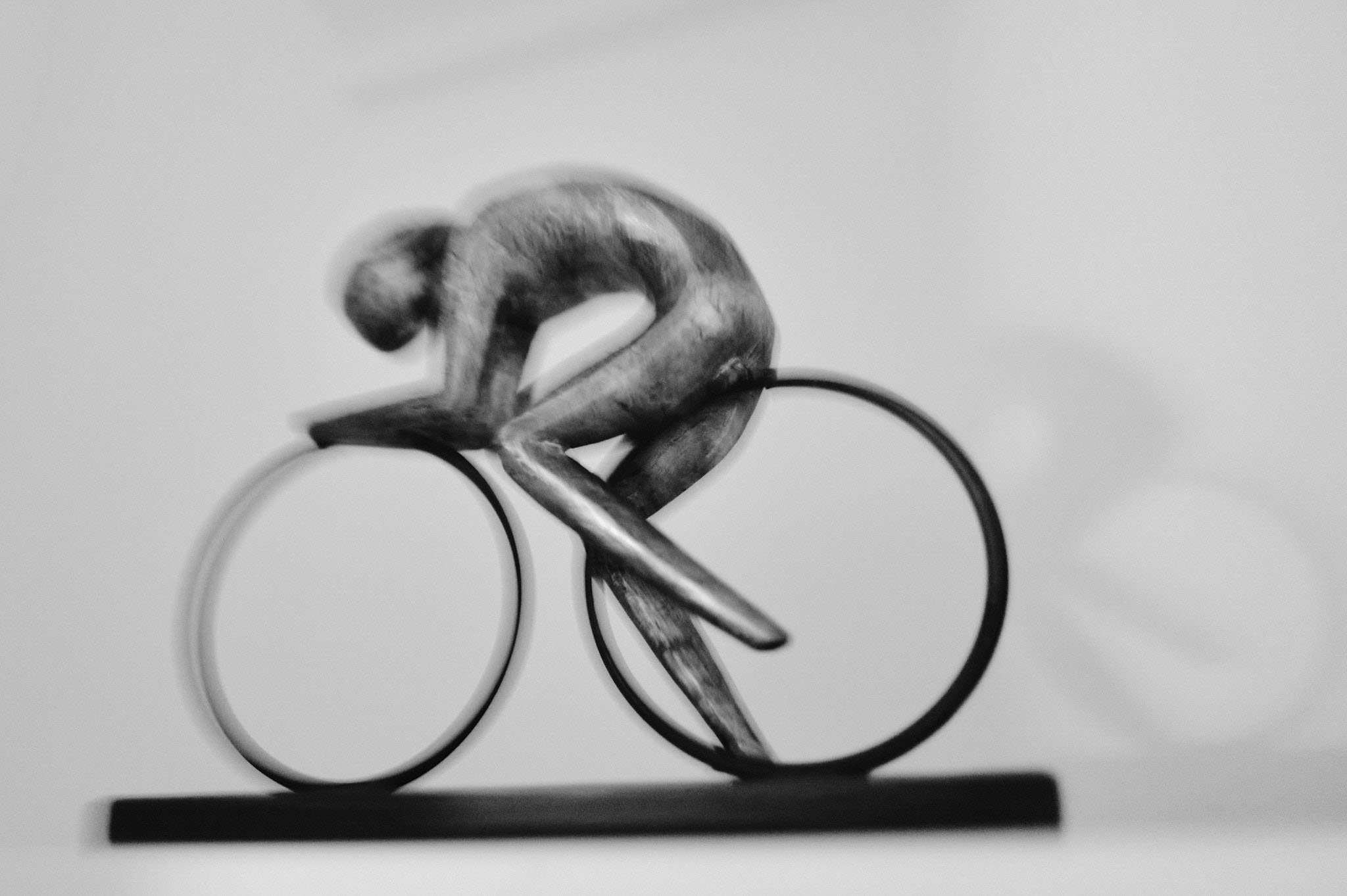 Black and white photograph of a pin-shaped bicycle sculpture with a figure riding it, mounted on a flat base.