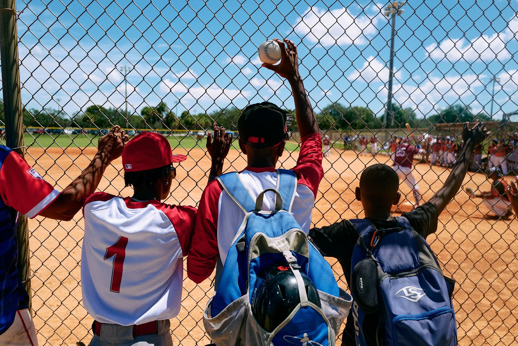 Three children with backpacks watching a baseball game through a chain-link fence on a sunny day.