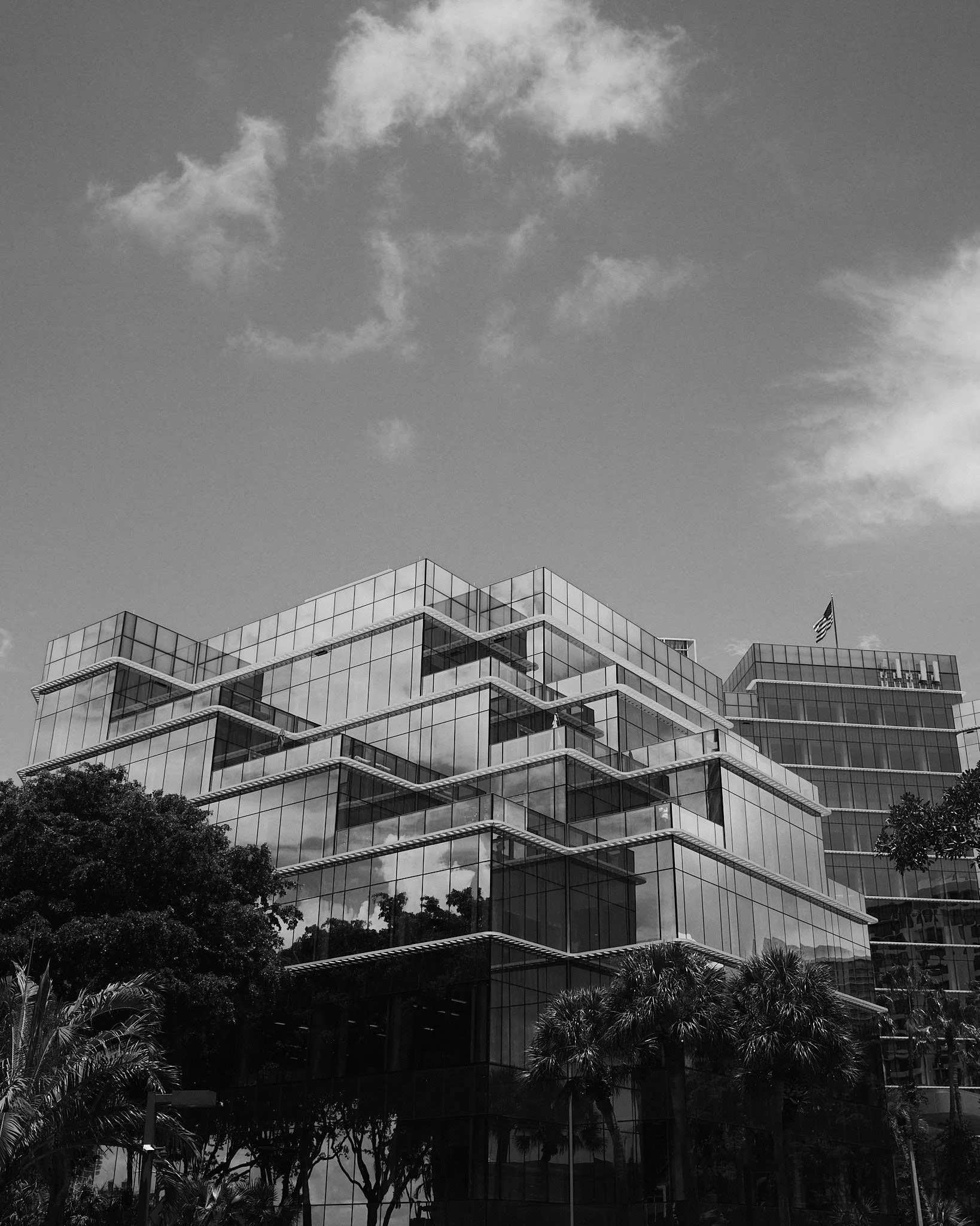 Modern glass office building with angular architecture and trees in front, under partly cloudy sky.
