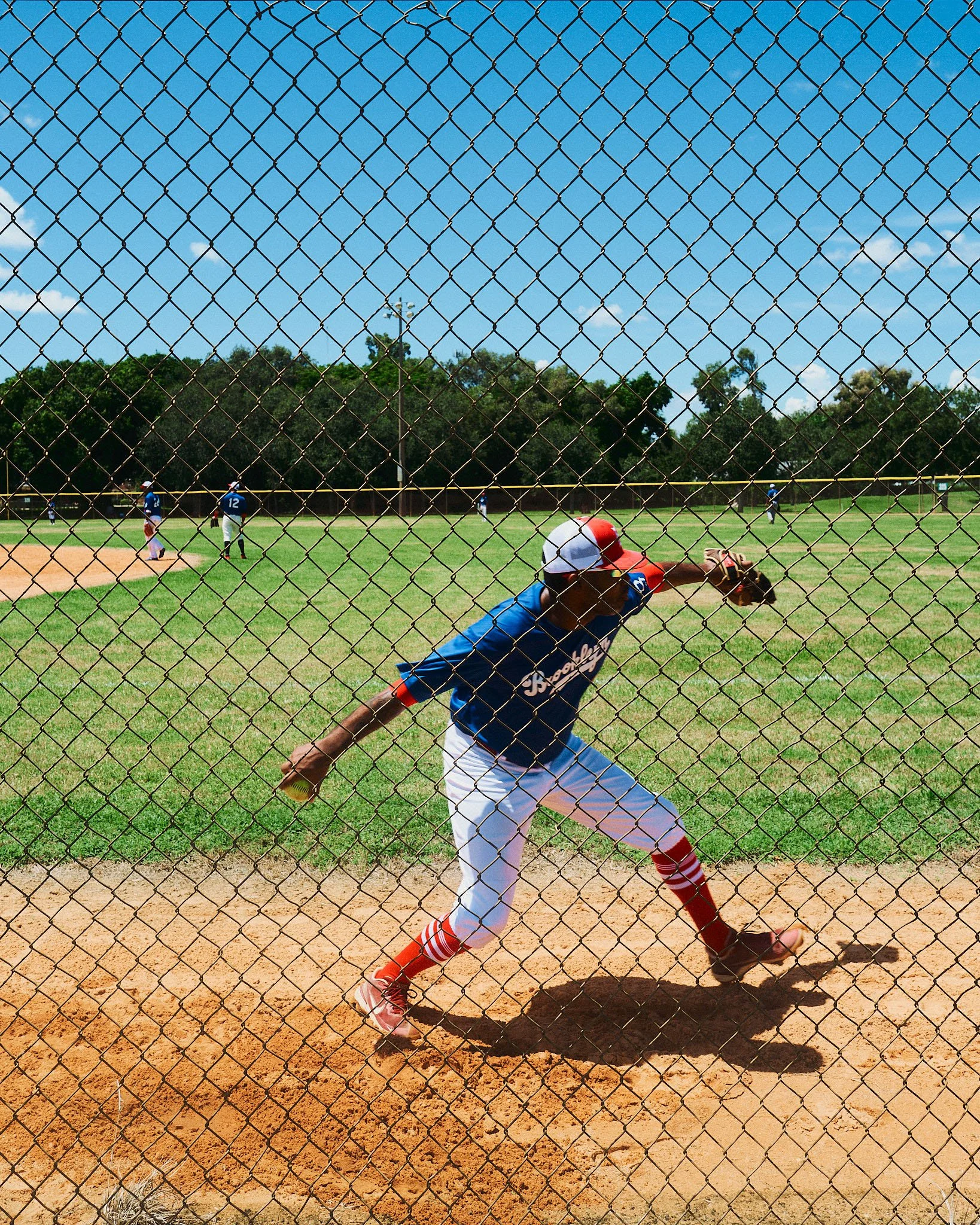 A baseball player wearing a blue jersey with white and red accents, white pants, red socks, and a red, white, and blue cap, throwing a baseball from the pitcher's mound on a baseball field. The game is in progress with other players visible in the ba