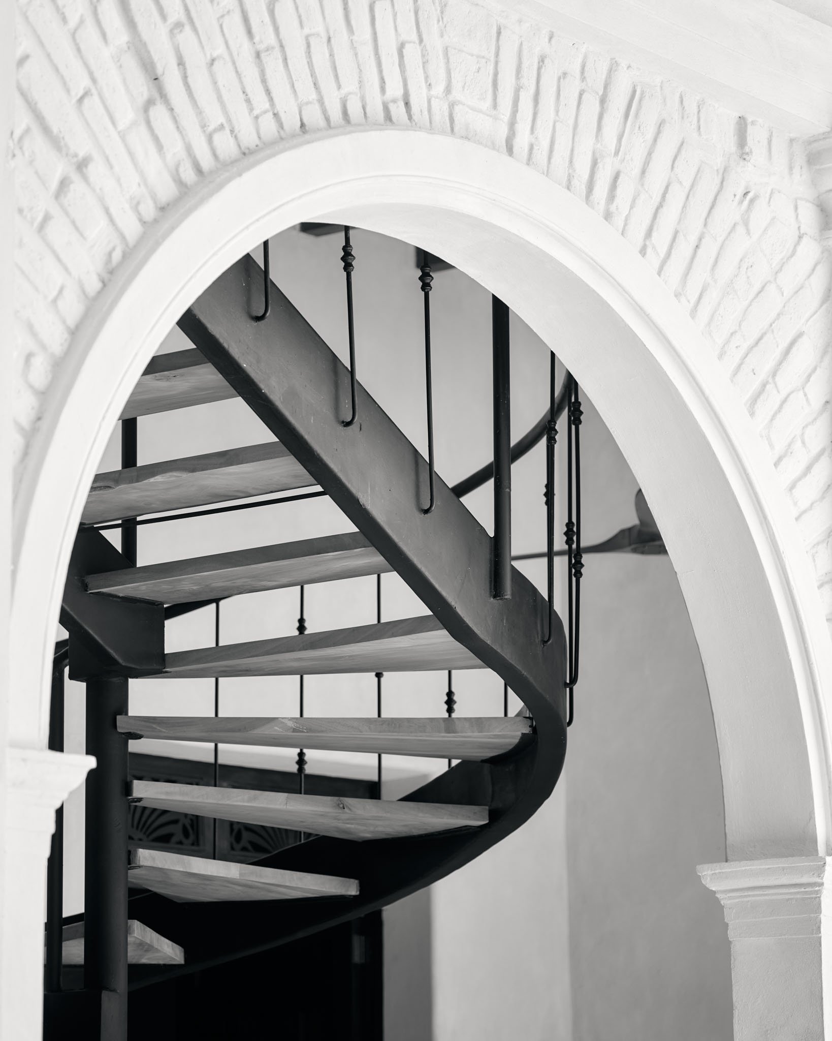 Black metal spiral staircase with wooden steps viewed through an arched white brick wall.