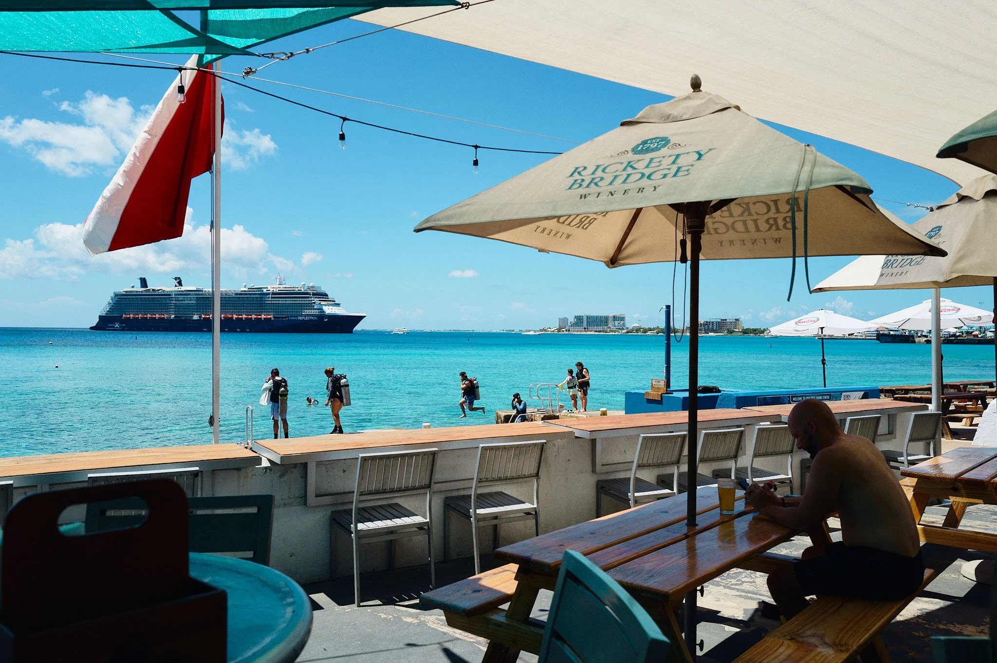 Beachside view with umbrellas and seating at a restaurant, people swimming and walking near the water, and a large cruise ship in the background on a sunny day.