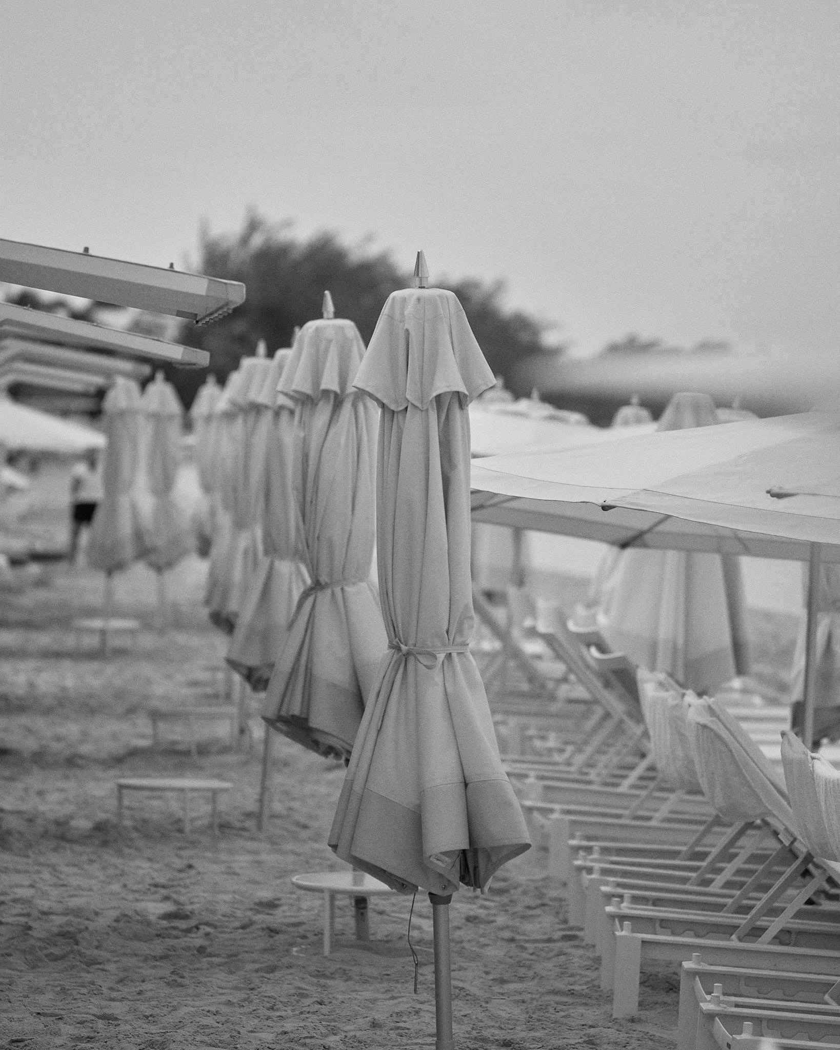 Row of closed beach umbrellas on a sandy beach, with lounge chairs underneath and a cloudy sky in the background.