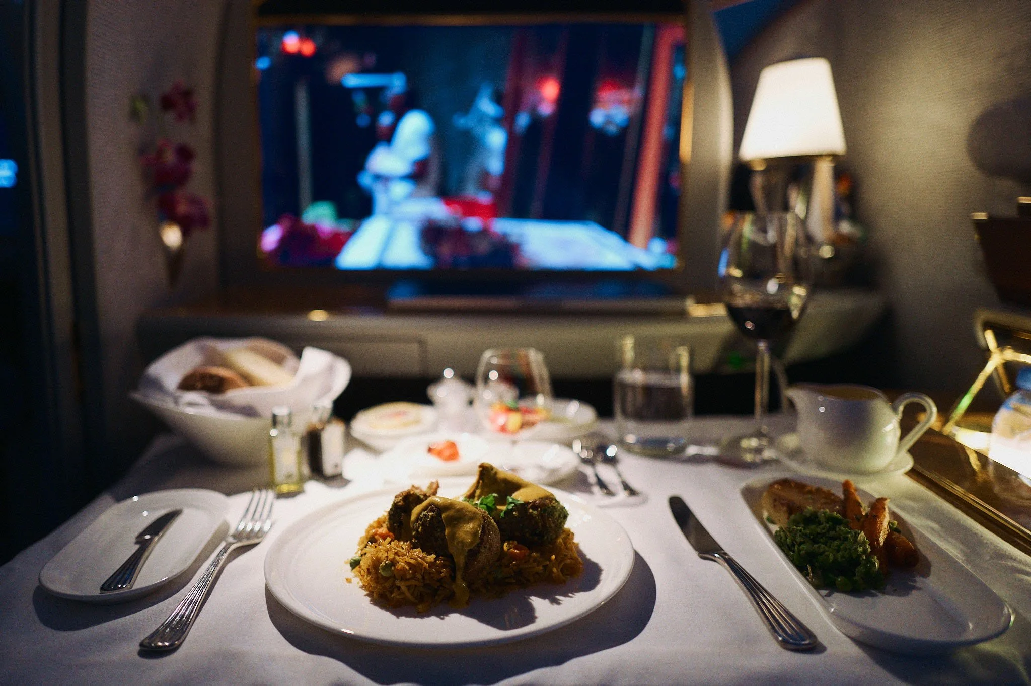 In-flight airline meal served on a tray table with a main dish of rice and meat topped with cheese, a side dish of vegetables, and bread in a basket. The background shows a blurred TV screen and a lamp, indicating a cozy setting inside a plane.