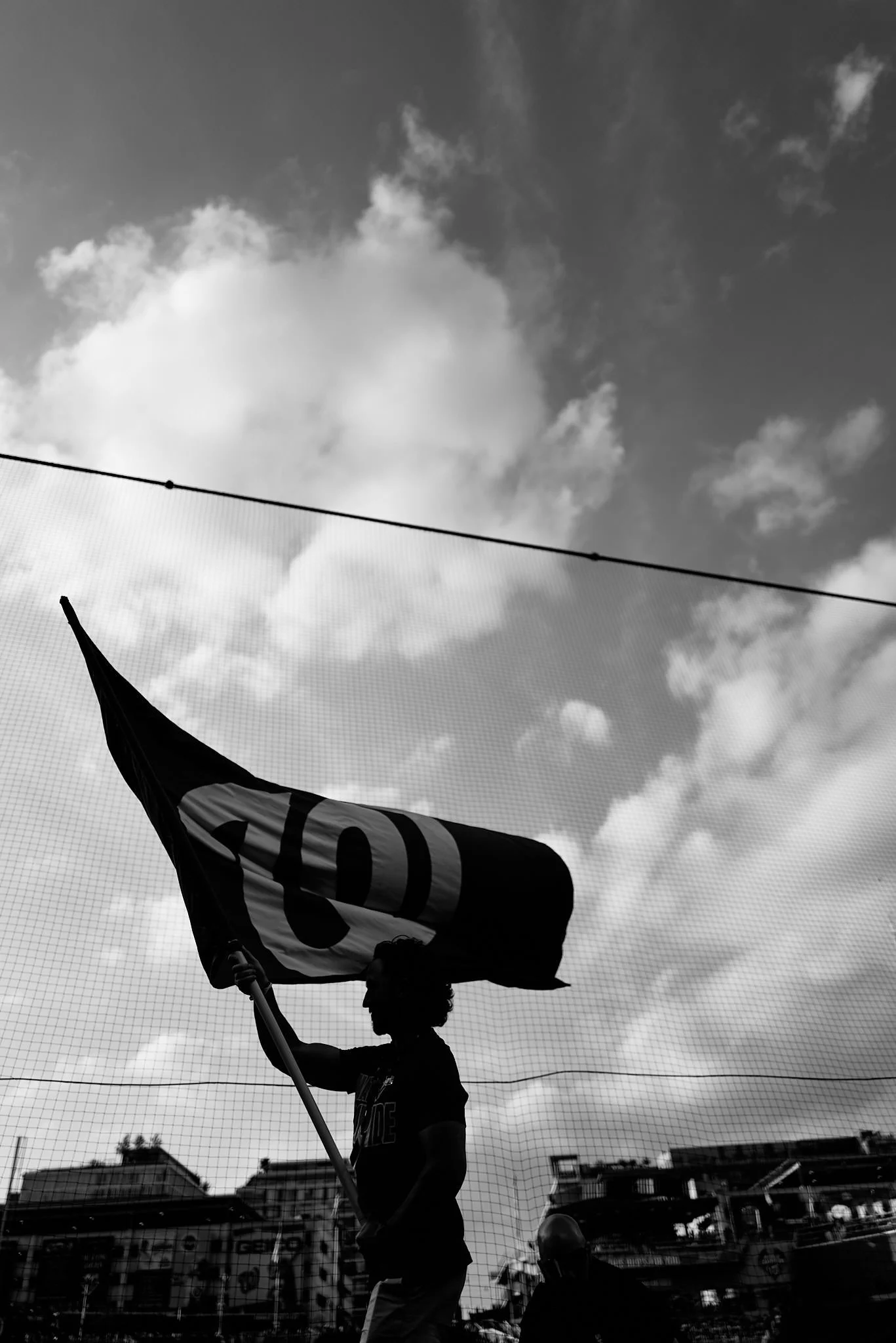A person holding a flag with the word 'STOP' on it, silhouette against a sky with clouds. There are buildings and a fence in the background.