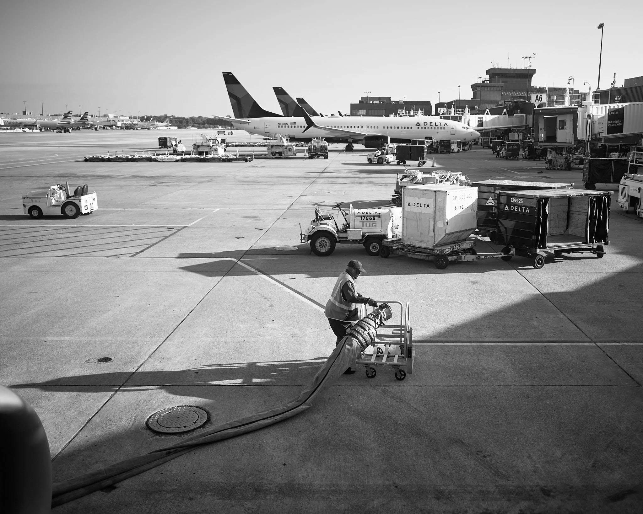 At an airport, ground crew member handling a large hose or cable near baggage carts, with airplanes and airport terminals in the background.