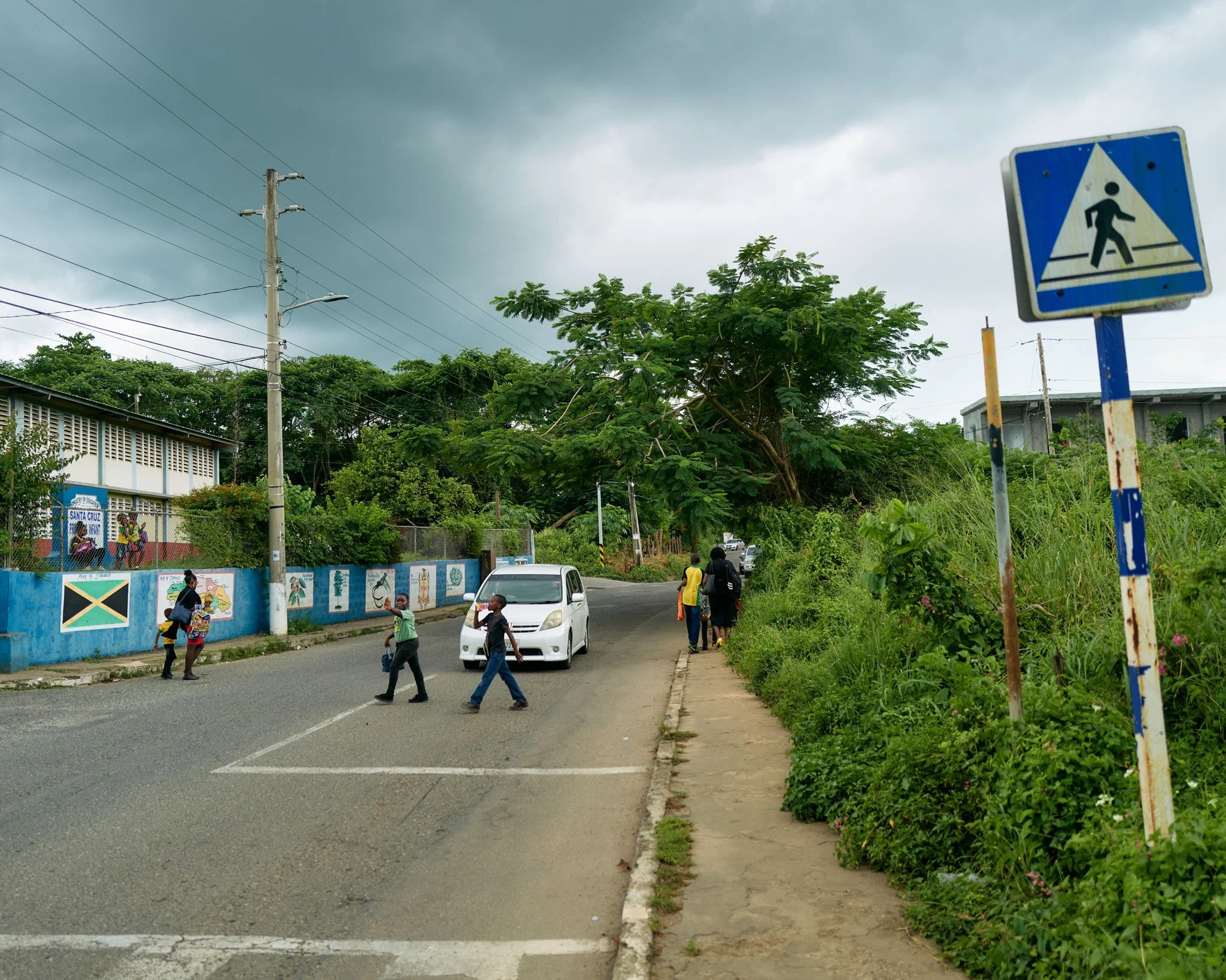 Children crossing the street near a school with a blue and white pedestrian crossing sign, and a partially cloudy sky.
