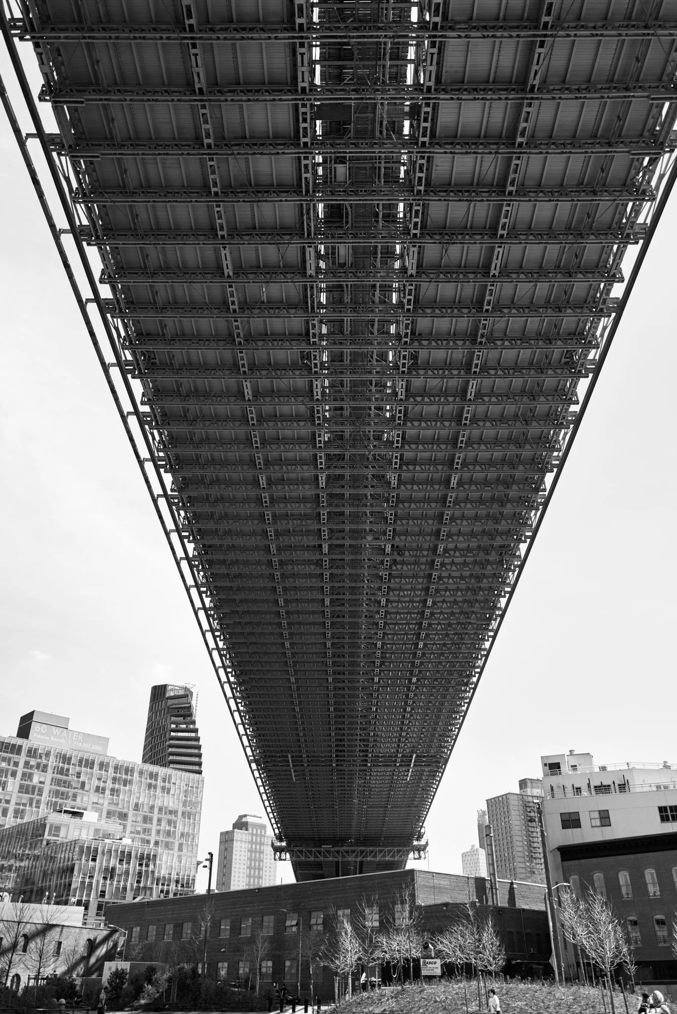 A low-angle view of a large steel bridge structure in an urban area, with tall buildings and skyscrapers in the background, black and white photograph.