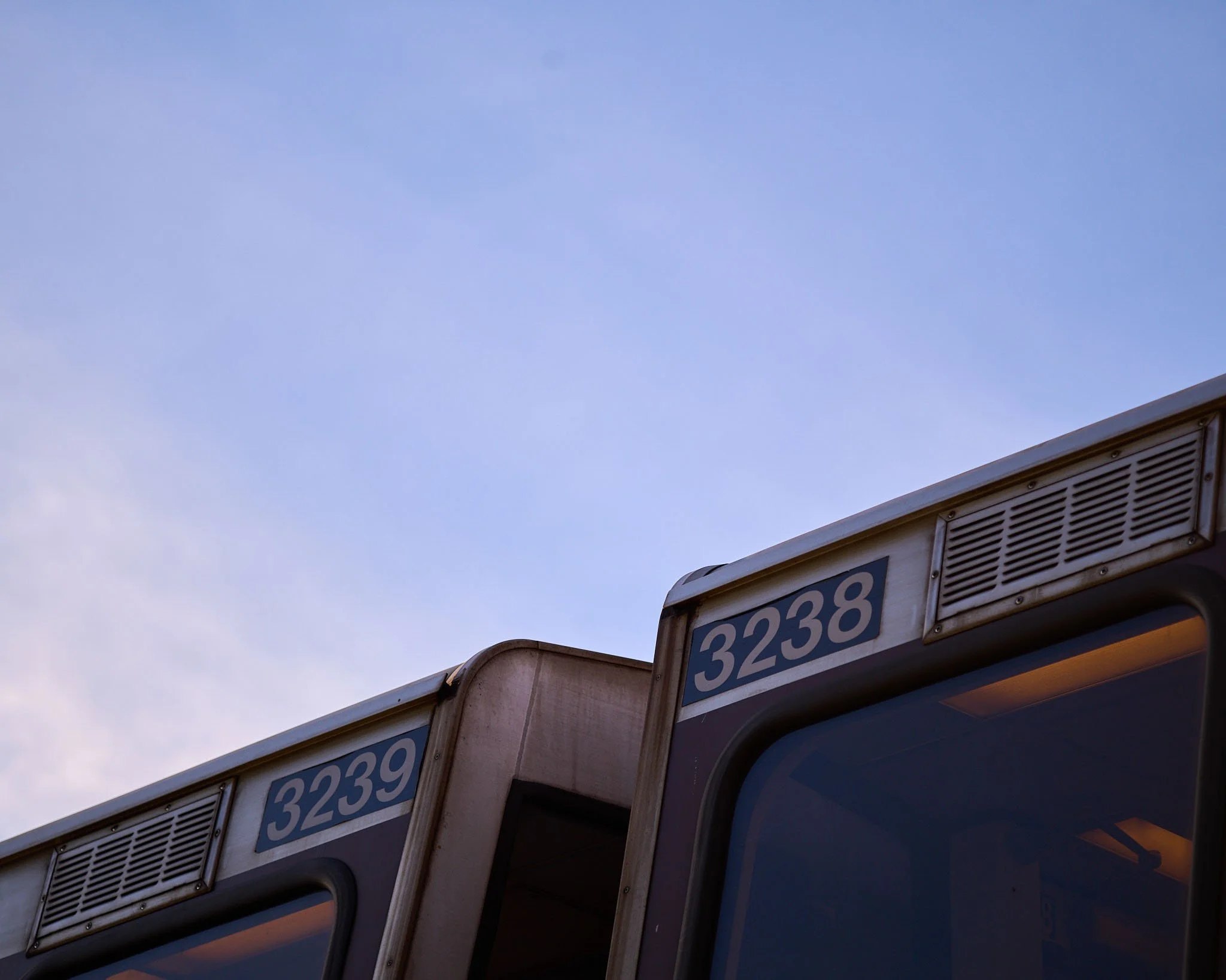 Close-up of the front part of two train cars with window and identification numbers 3238 and 3239, against a partly cloudy sky.
