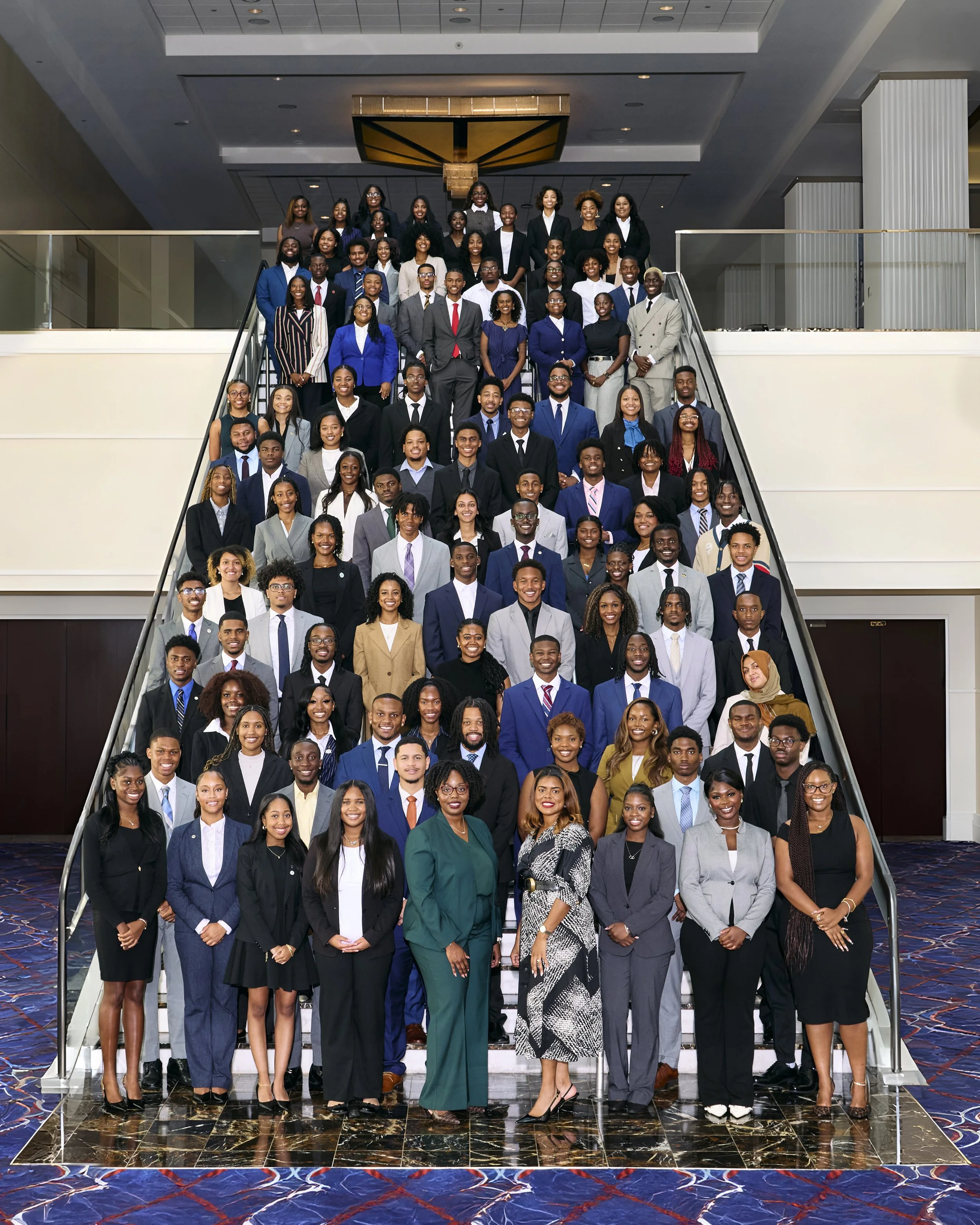 Group of diverse young professionals dressed in formal business attire, posing on a grand staircase in a luxurious hotel or conference center.
