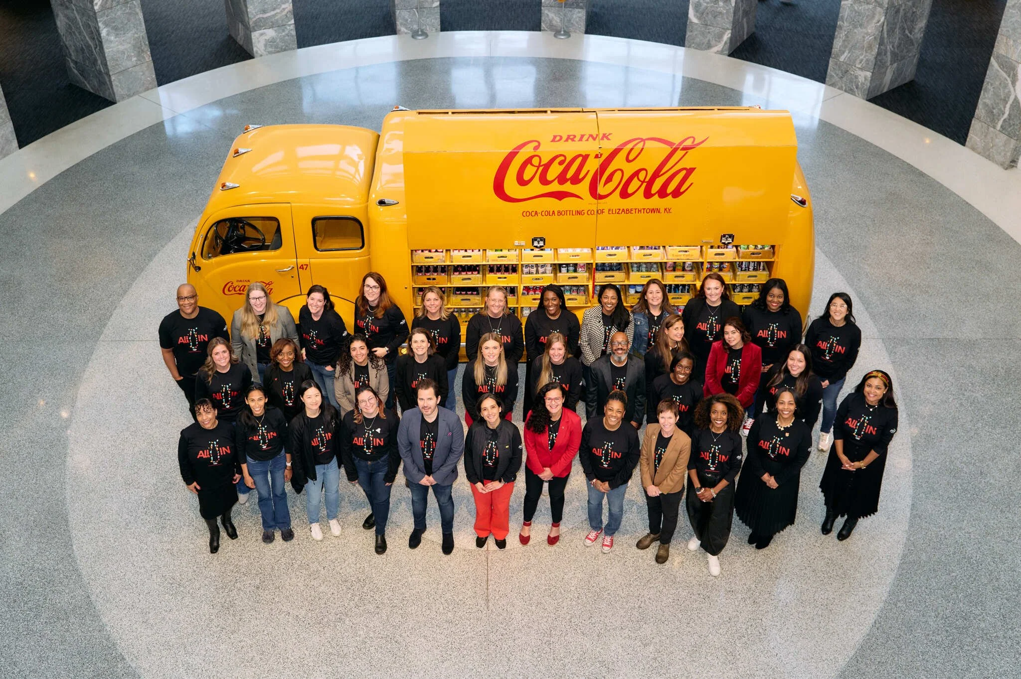 A group of diverse people standing in front of a large yellow Coca-Cola delivery truck inside a spacious, modern building with a circular gray and white patterned floor.